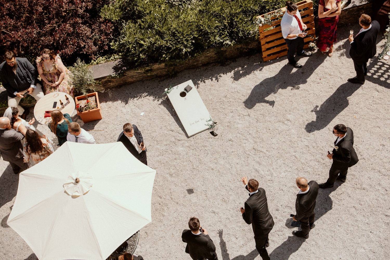 Aerial view of a wedding cocktail hour with guests mingling, cornhole game, and patio umbrella in a sunny courtyard.
