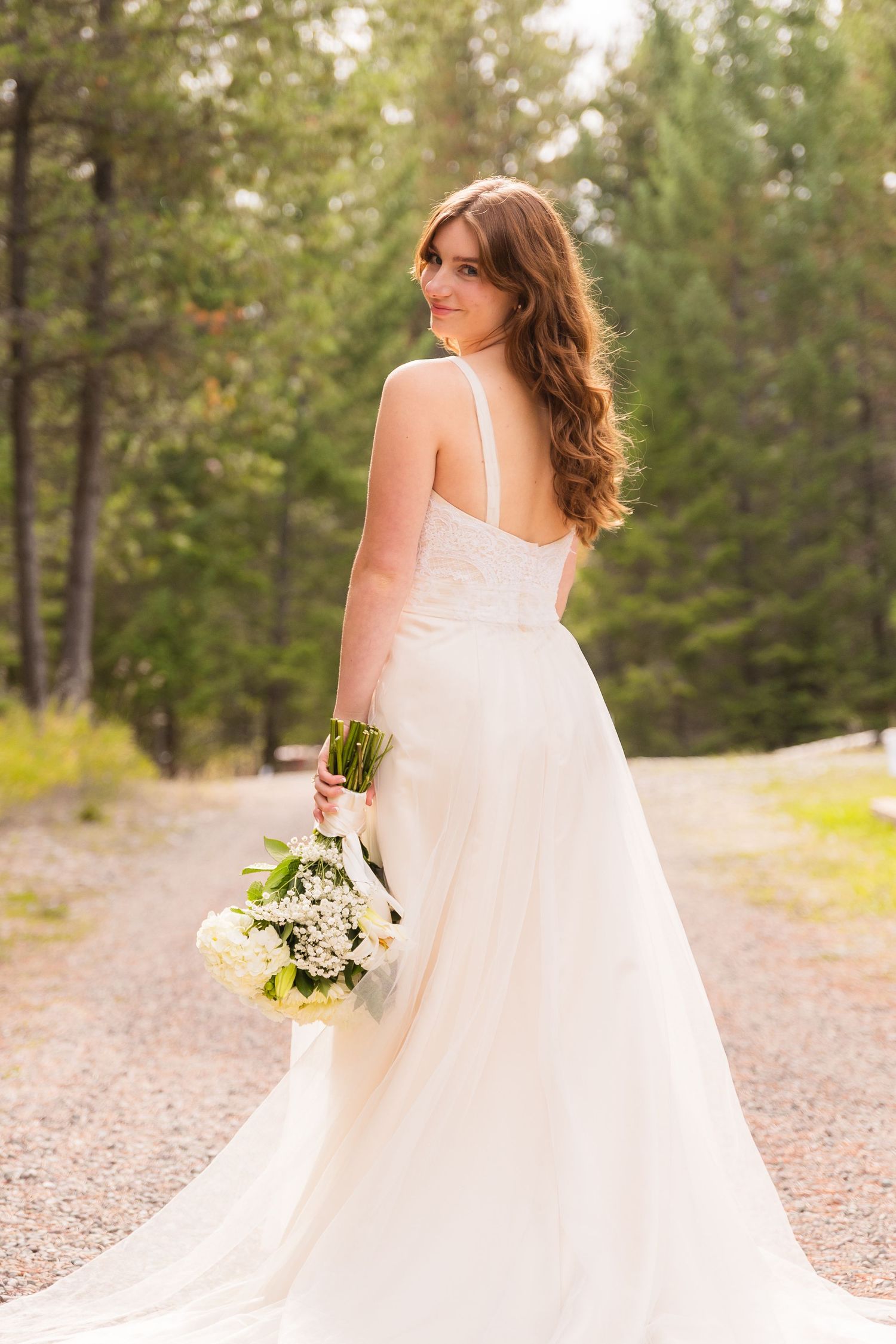 Bride in white gown holding bouquet on forest path.