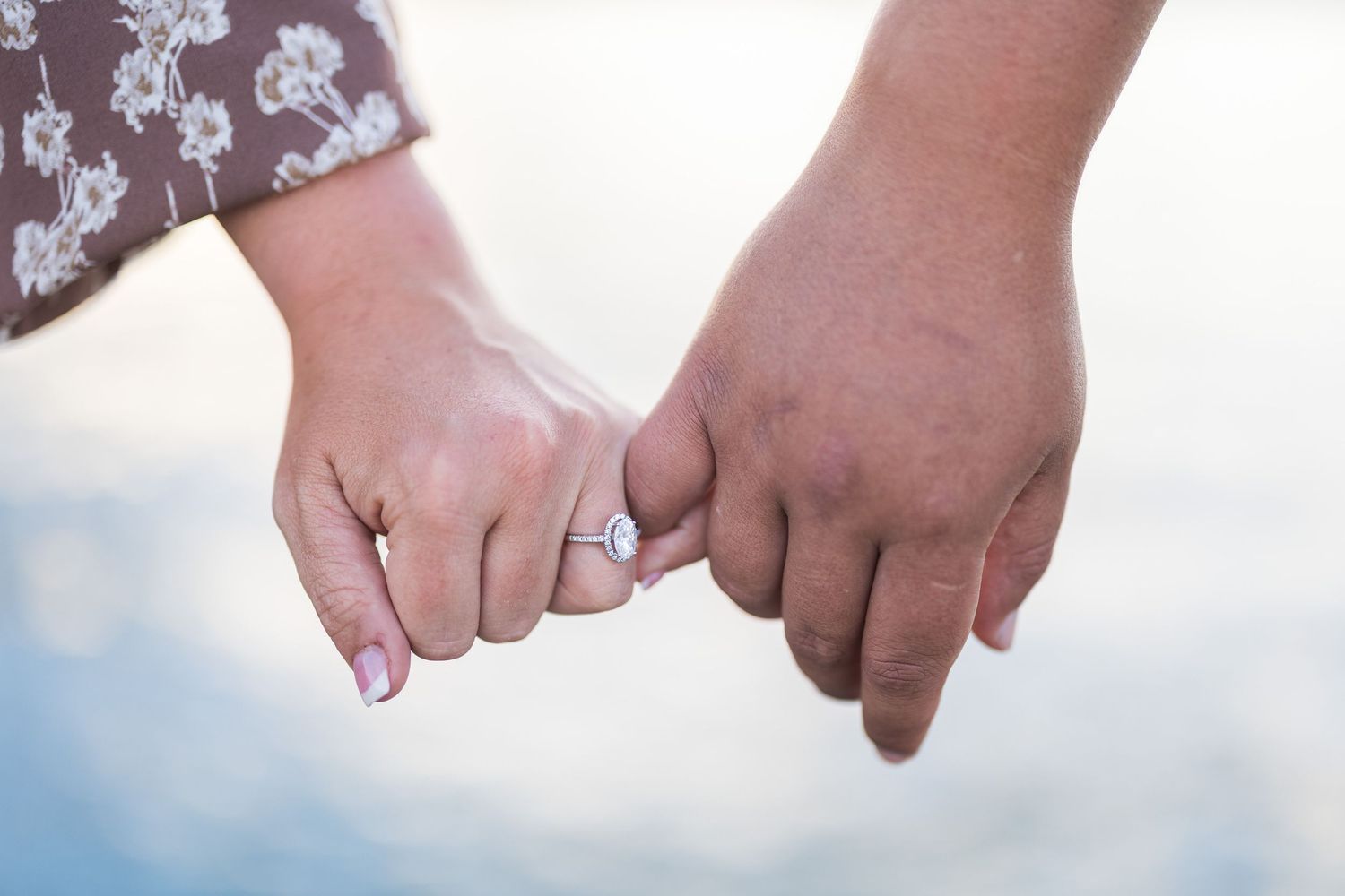 Close-up of couple holding pinkies with engagement ring visible against soft blurred background.
