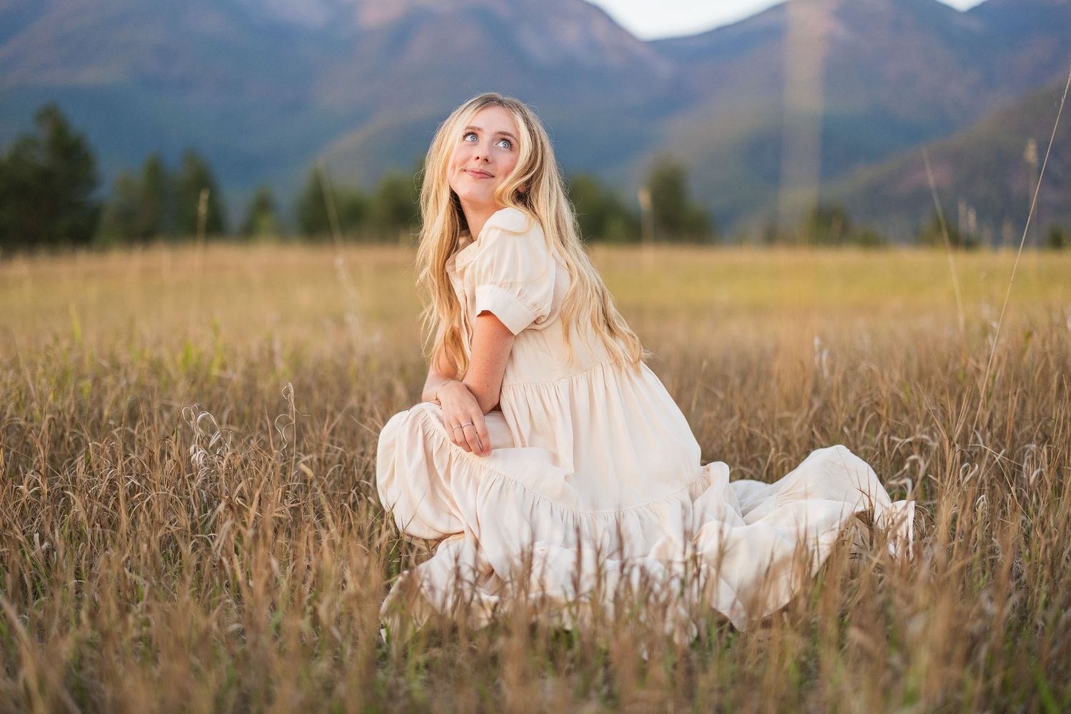 Blonde woman in white dress kneeling in a golden field with mountains in the background.
