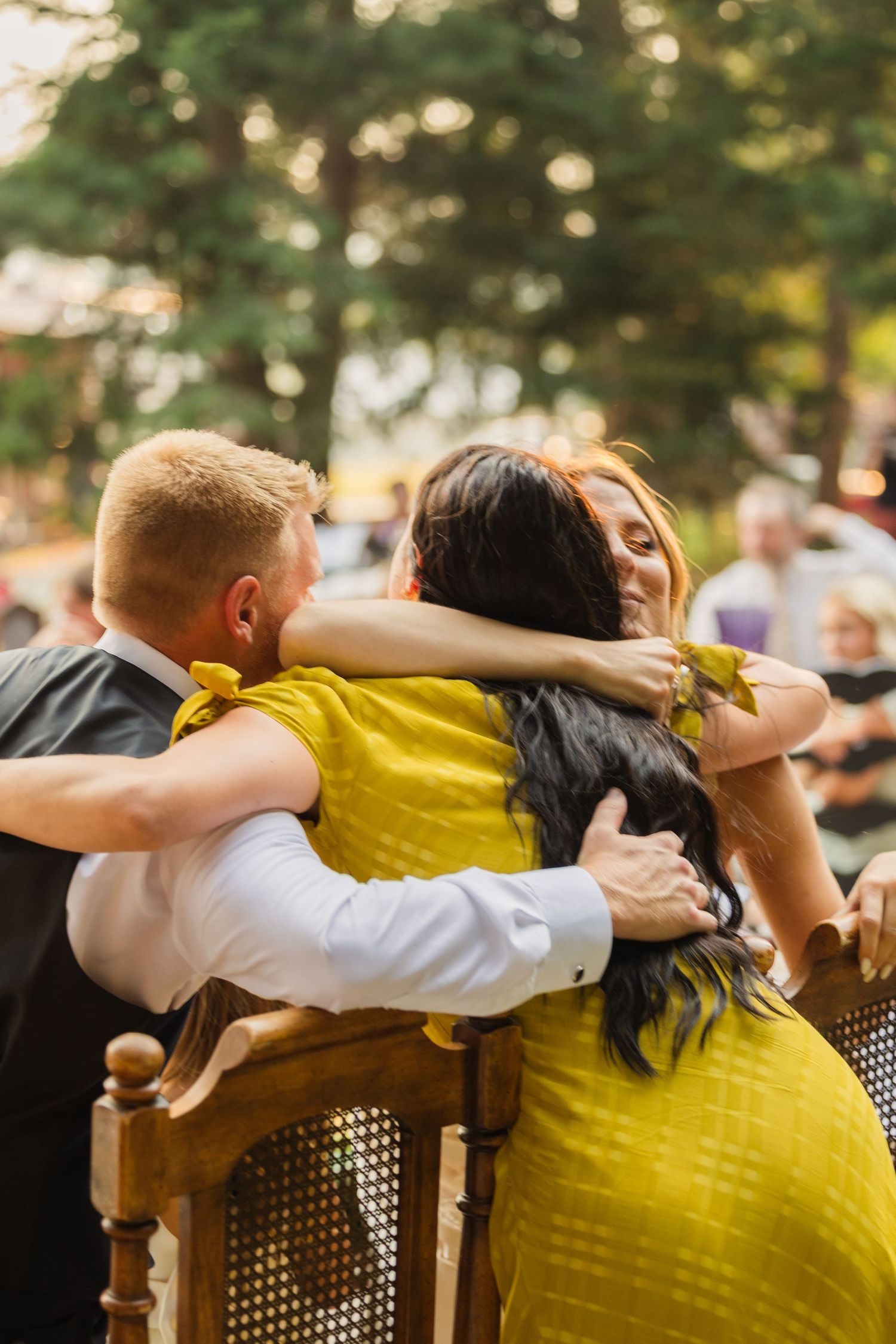 Three wedding guests share a warm group hug seated outdoors, two wearing yellow attire at reception.