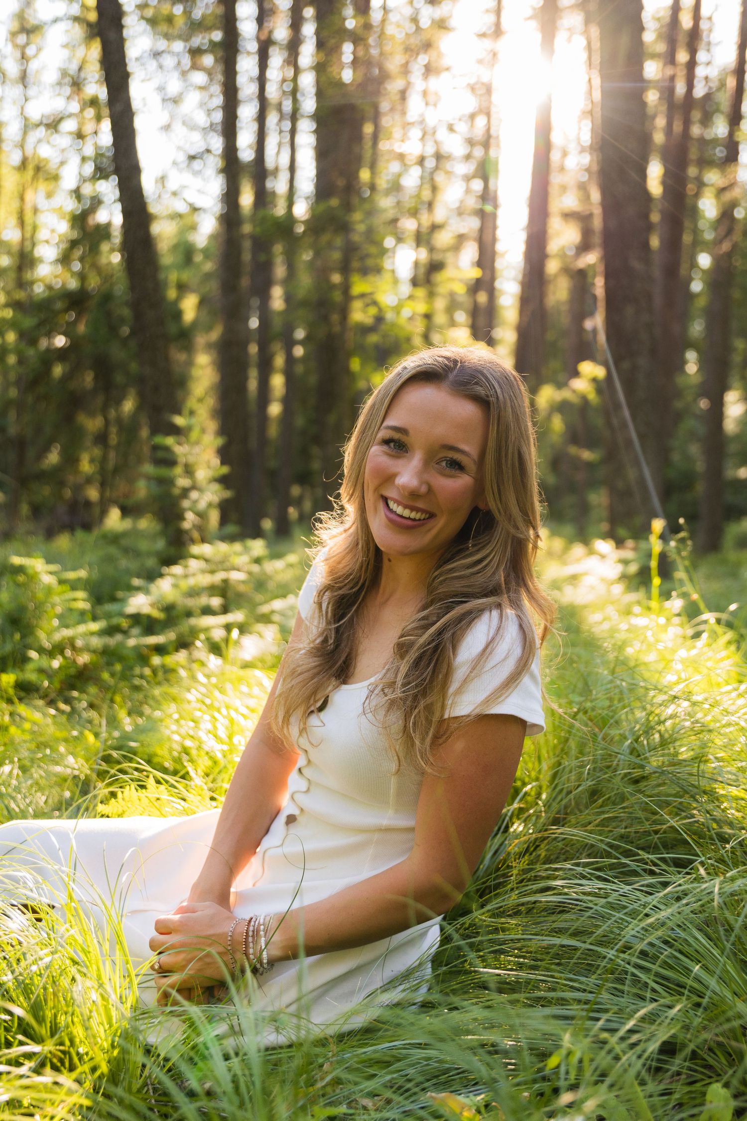 Smiling young woman in white dress sitting in lush green forest grass with golden sunlight filtering through trees.