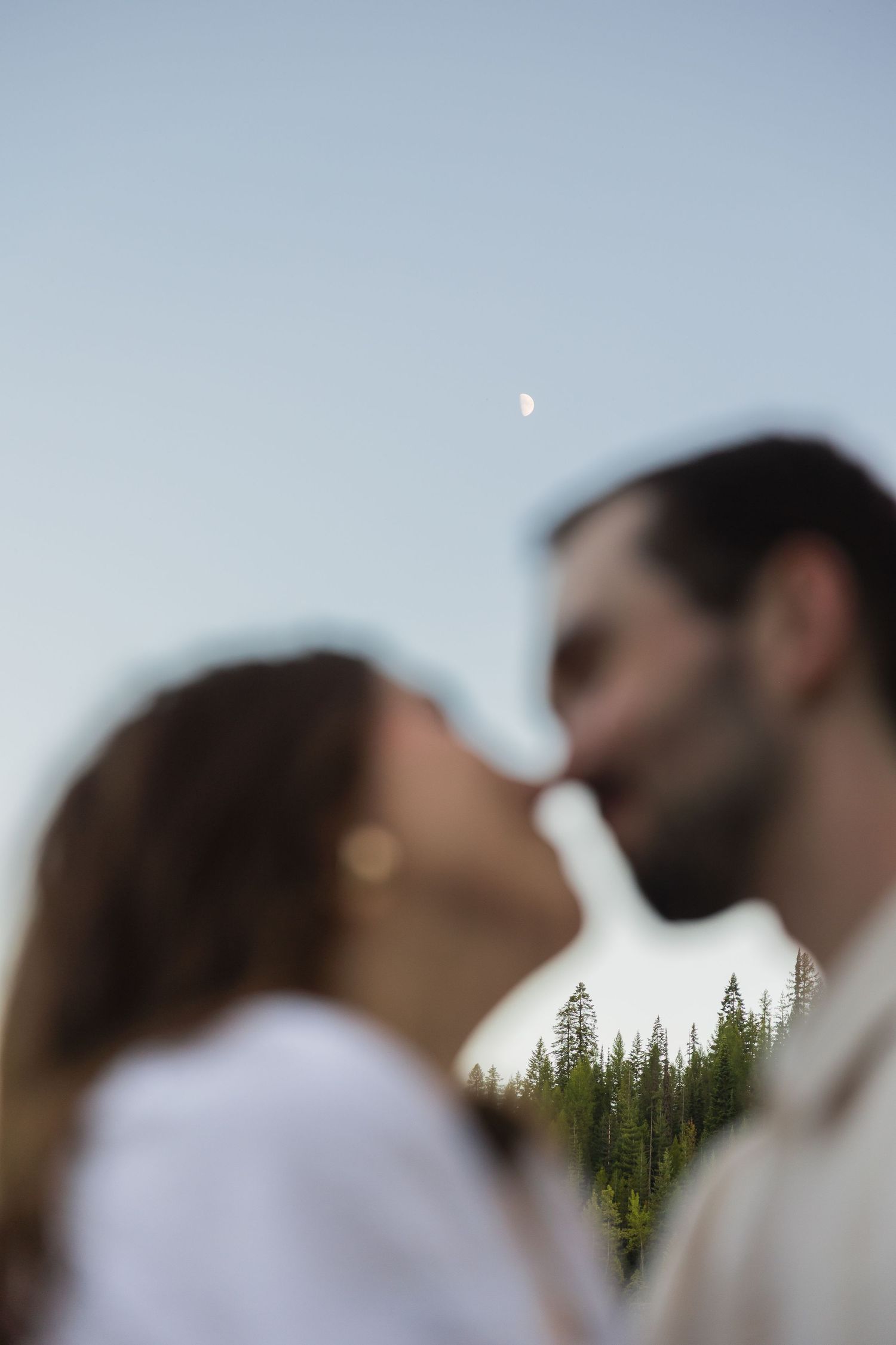 Romantic couple kissing outdoors with evergreen trees and a crescent moon visible in the soft evening sky.