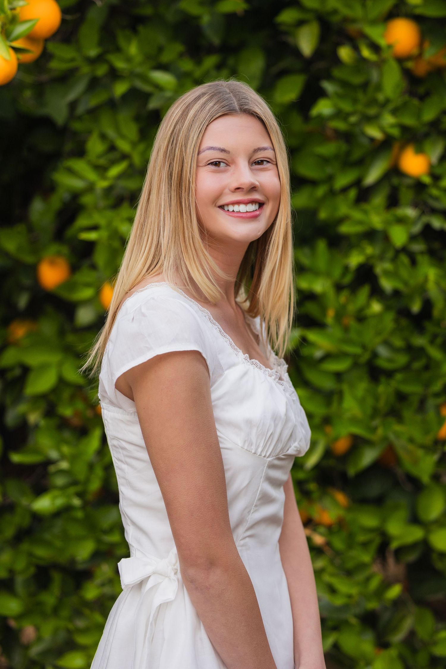 Smiling blonde teenage girl in white dress posing in front of an orange tree.
