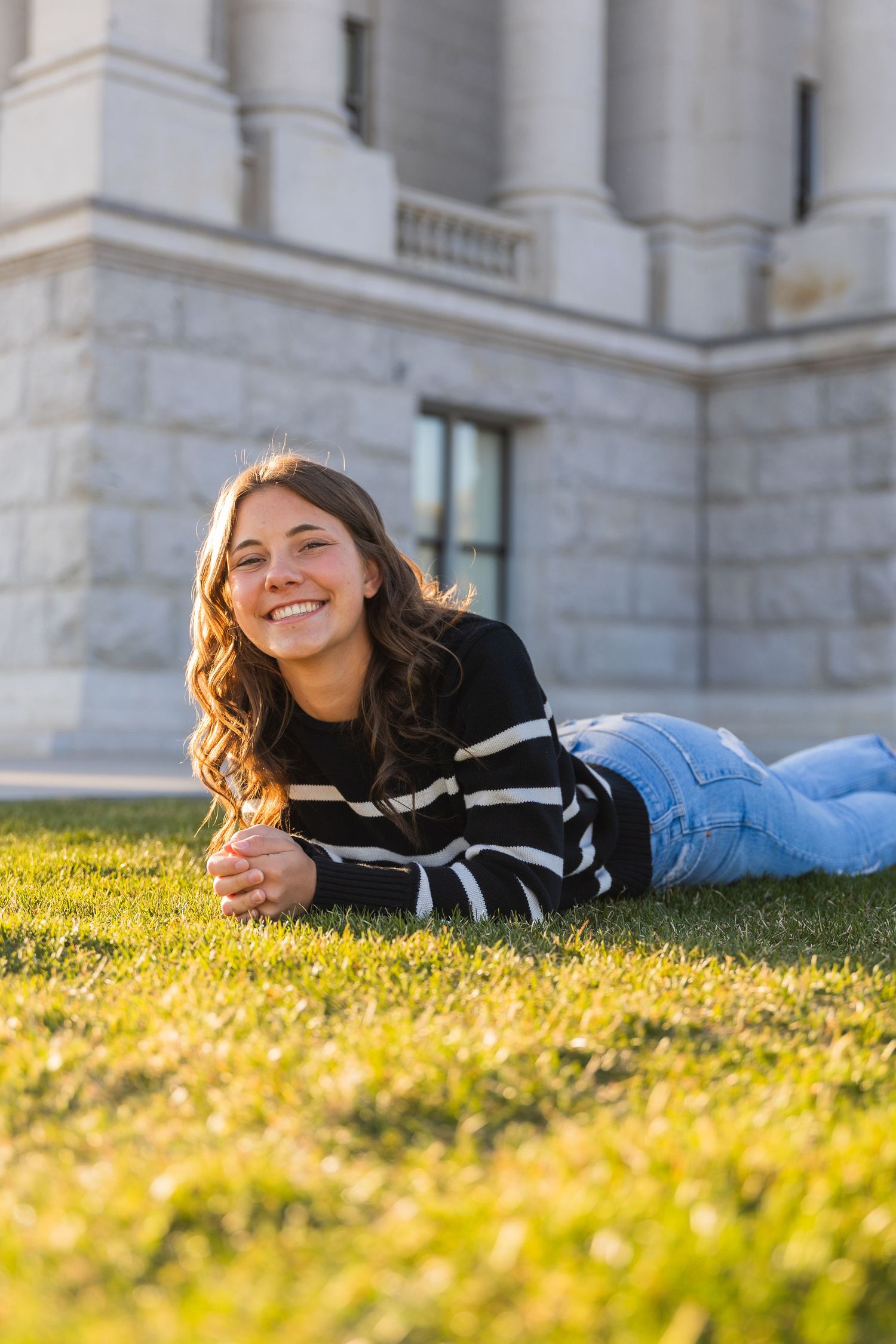 Smiling young woman lying on grass in front of a stone building in golden sunlight.