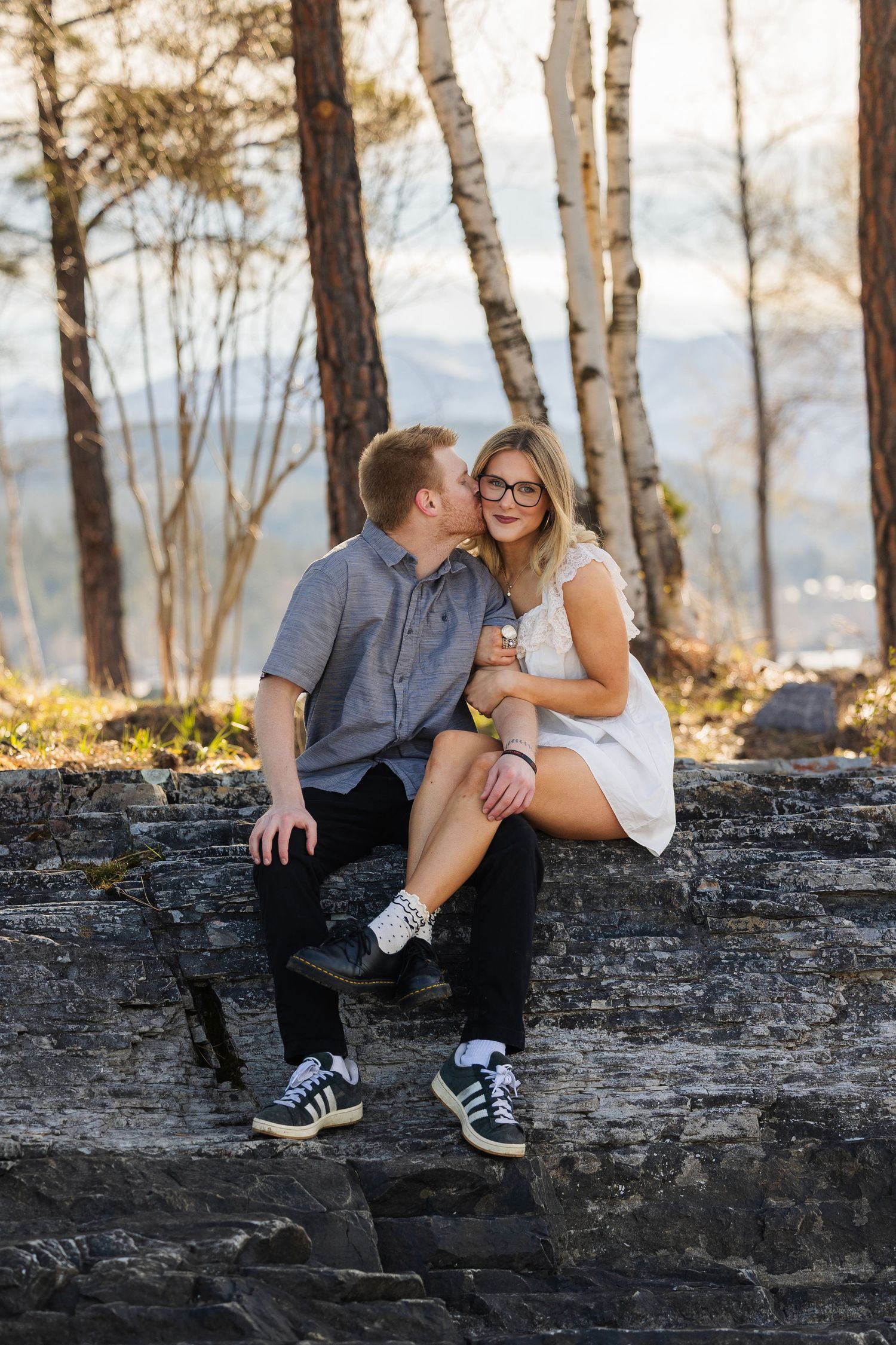 Young couple sitting intimately on rocky outcrop surrounded by birch trees in golden light.