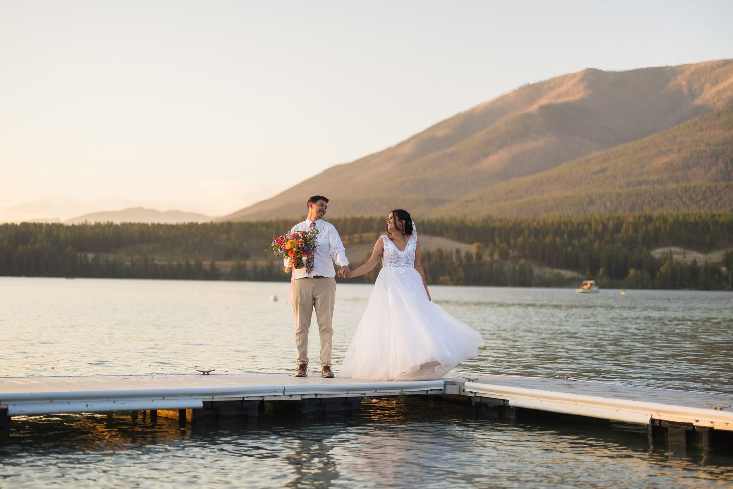 Bride and groom hold hands on a dock with a stunning mountain lake backdrop at sunset.