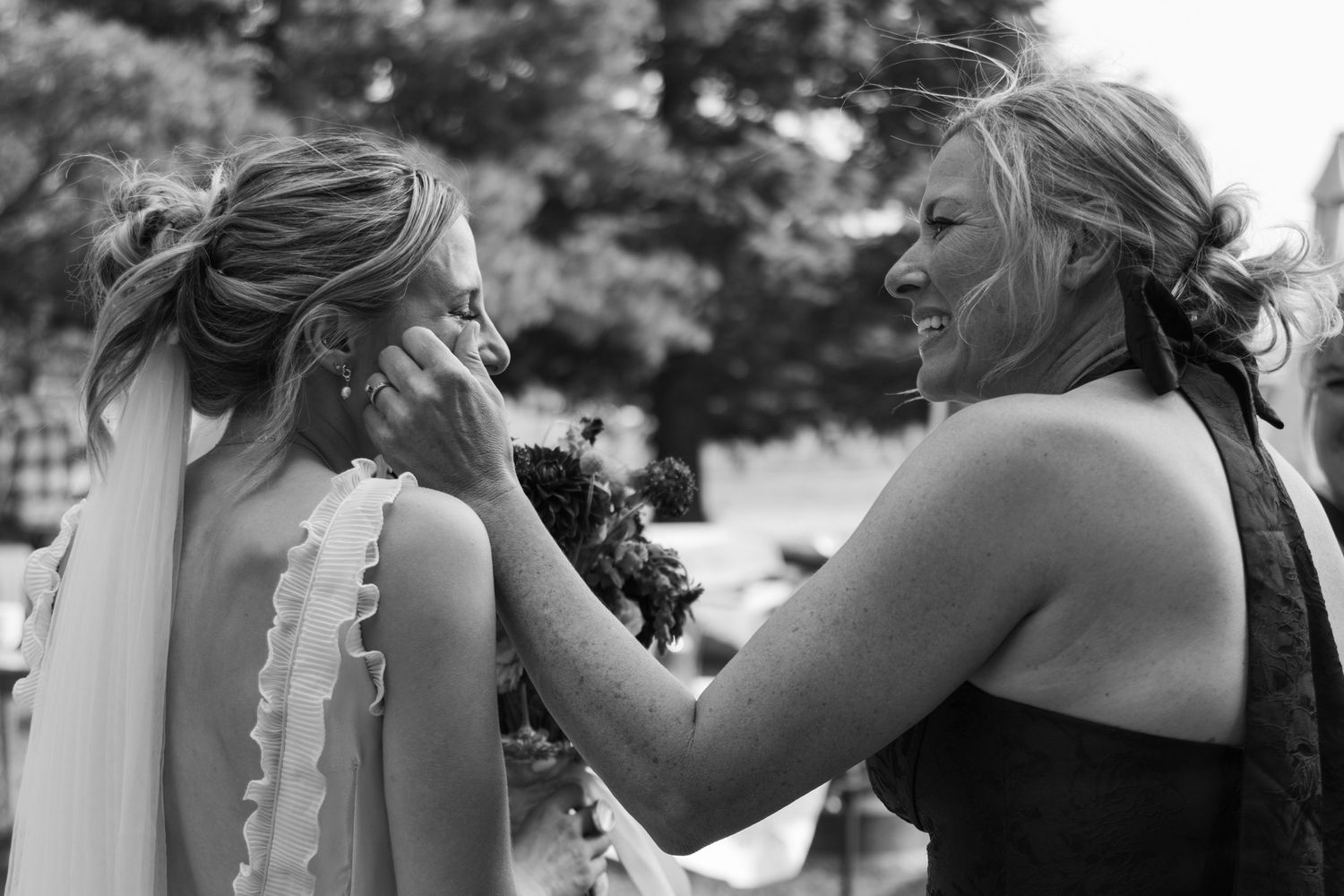 Bridesmaid tenderly wiping tears from bride's cheek in an emotional black and white wedding moment outdoors.