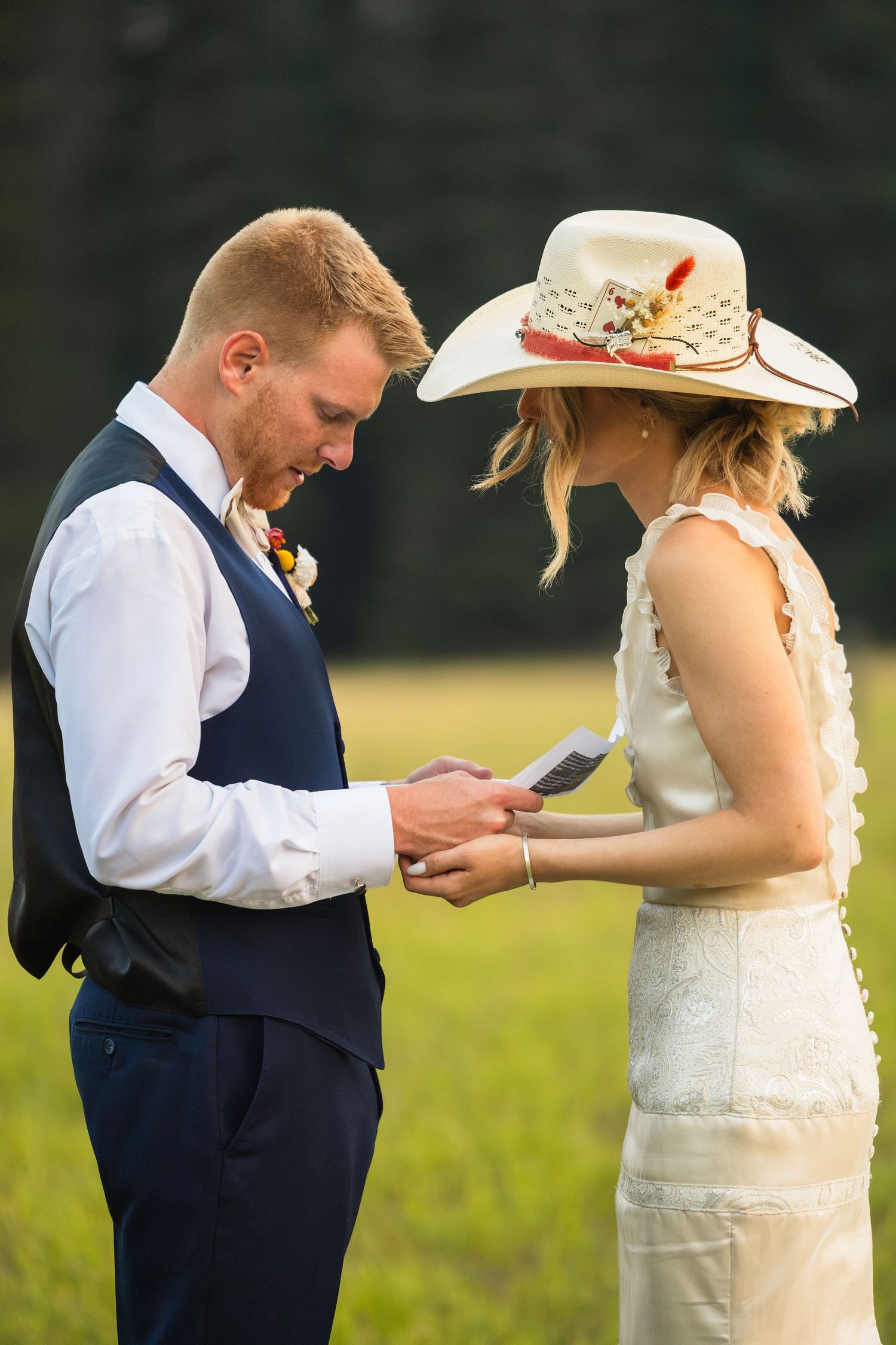 Groom in navy vest reads vows while bride in white dress and cowboy hat listens during outdoor wedding ceremony.