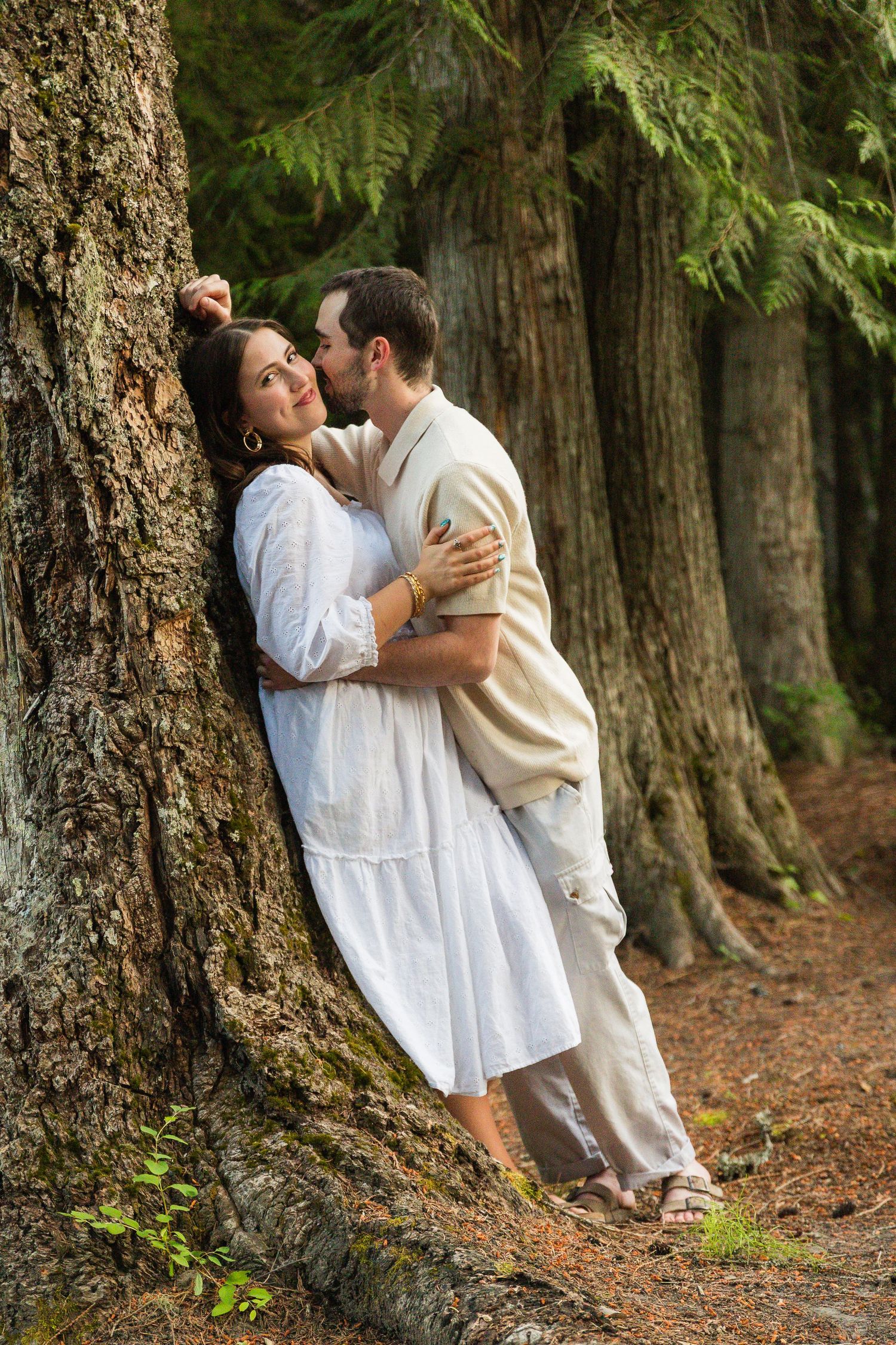 Romantic couple embracing by a large tree in a lush forest, woman in white dress, man in beige outfit.