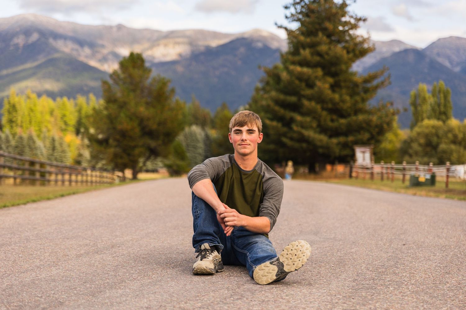 Young man sitting cross-legged on a road with mountains and trees in the background.
