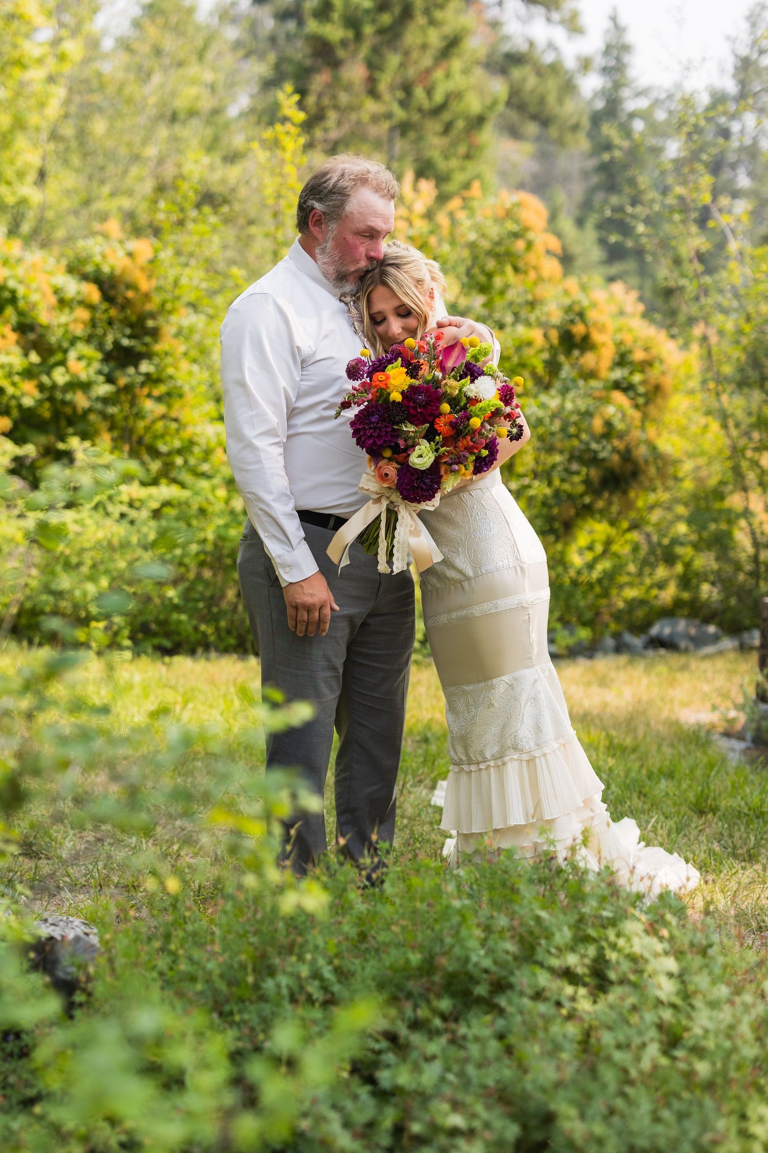 Bride and groom embrace outdoors among autumn foliage, bride holding vibrant red and yellow wedding bouquet.
