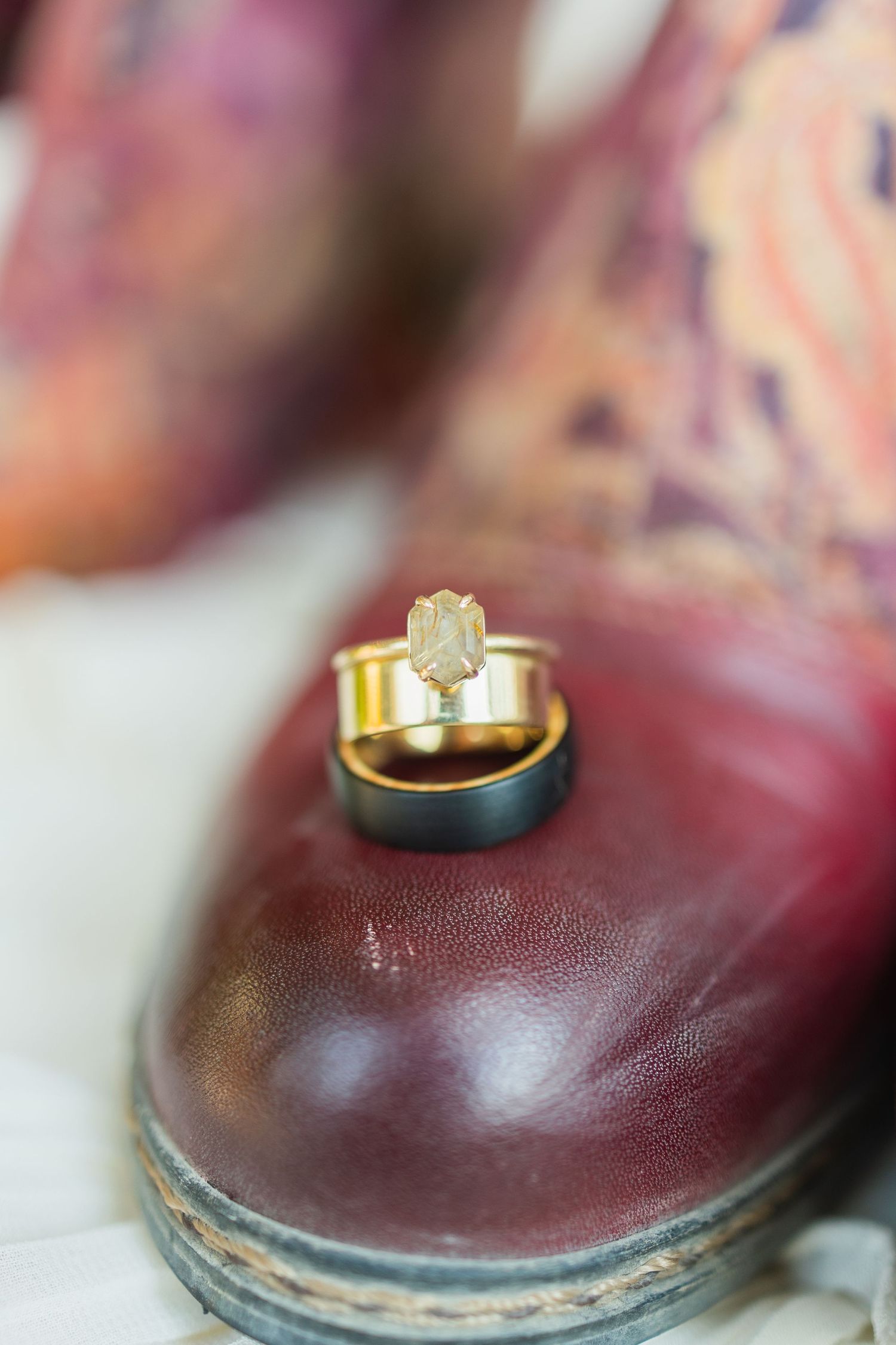 Gold engagement ring with raw diamond stone stacked on wedding bands displayed on a burgundy leather dress shoe.