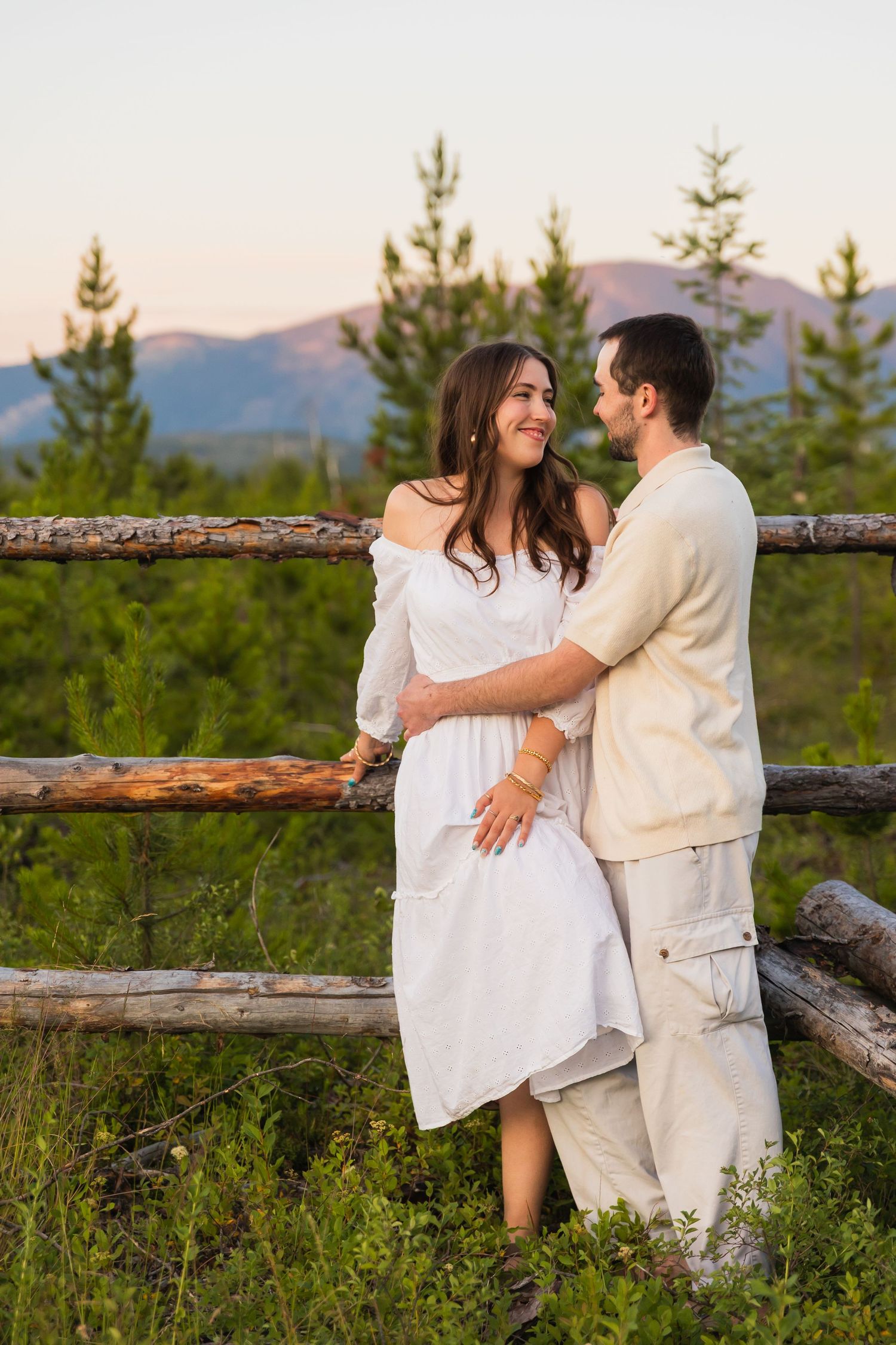 A couple embraces by a rustic log fence at sunset, surrounded by pine trees and mountain views.
