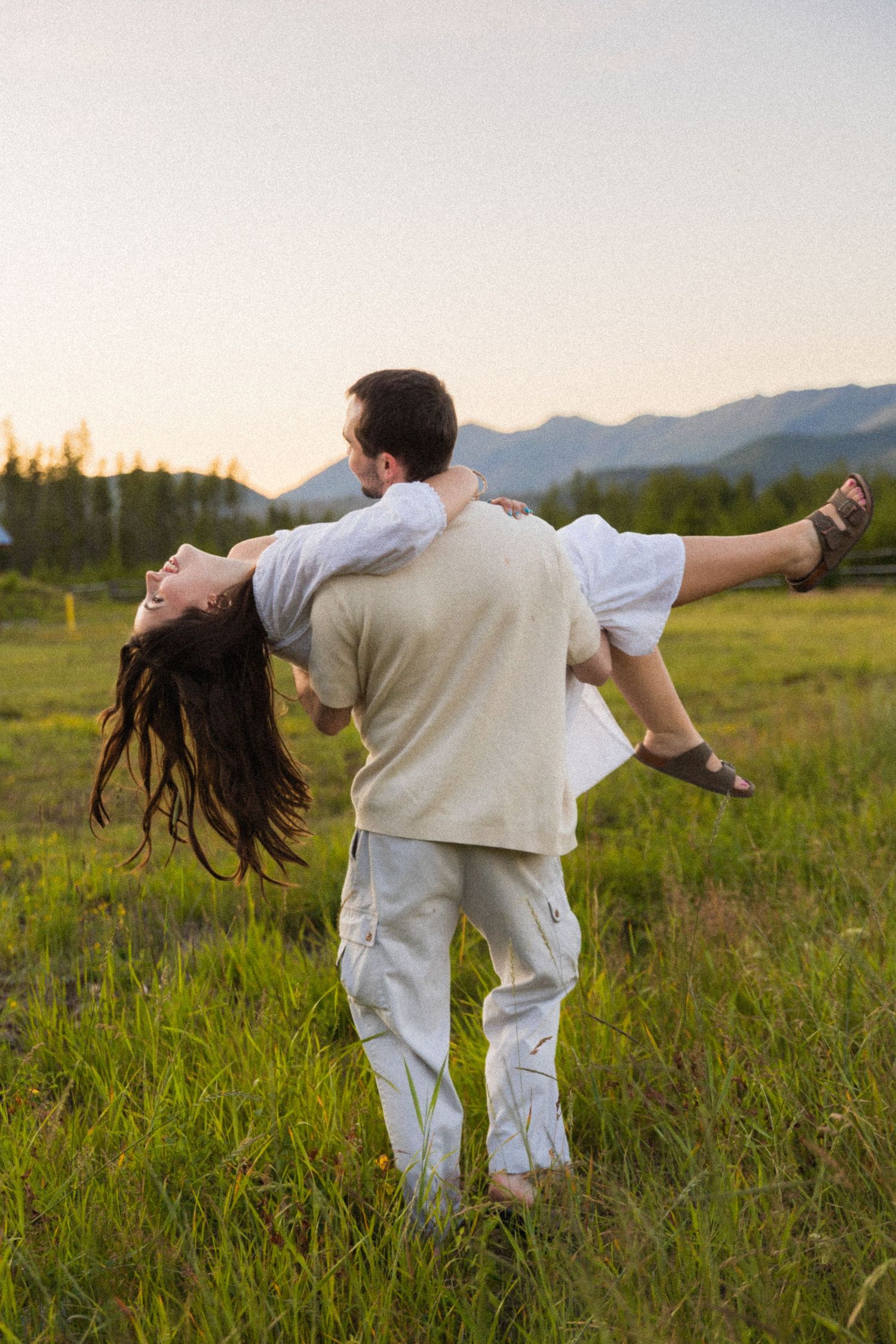 A man joyfully lifts and spins a woman in a green meadow with mountains at sunset.