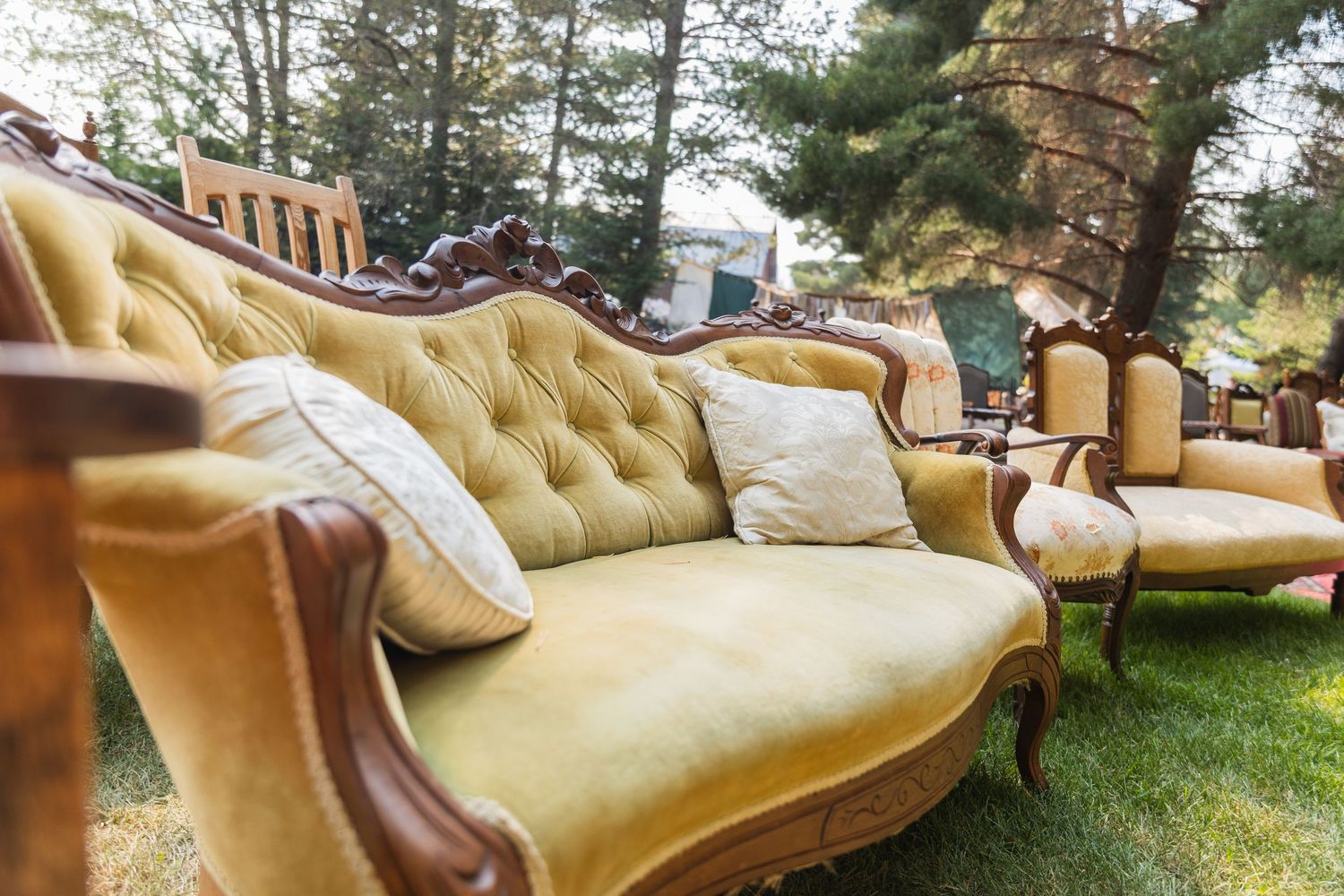 Vintage yellow tufted velvet sofa with ornate wooden frame displayed outdoors on green grass.