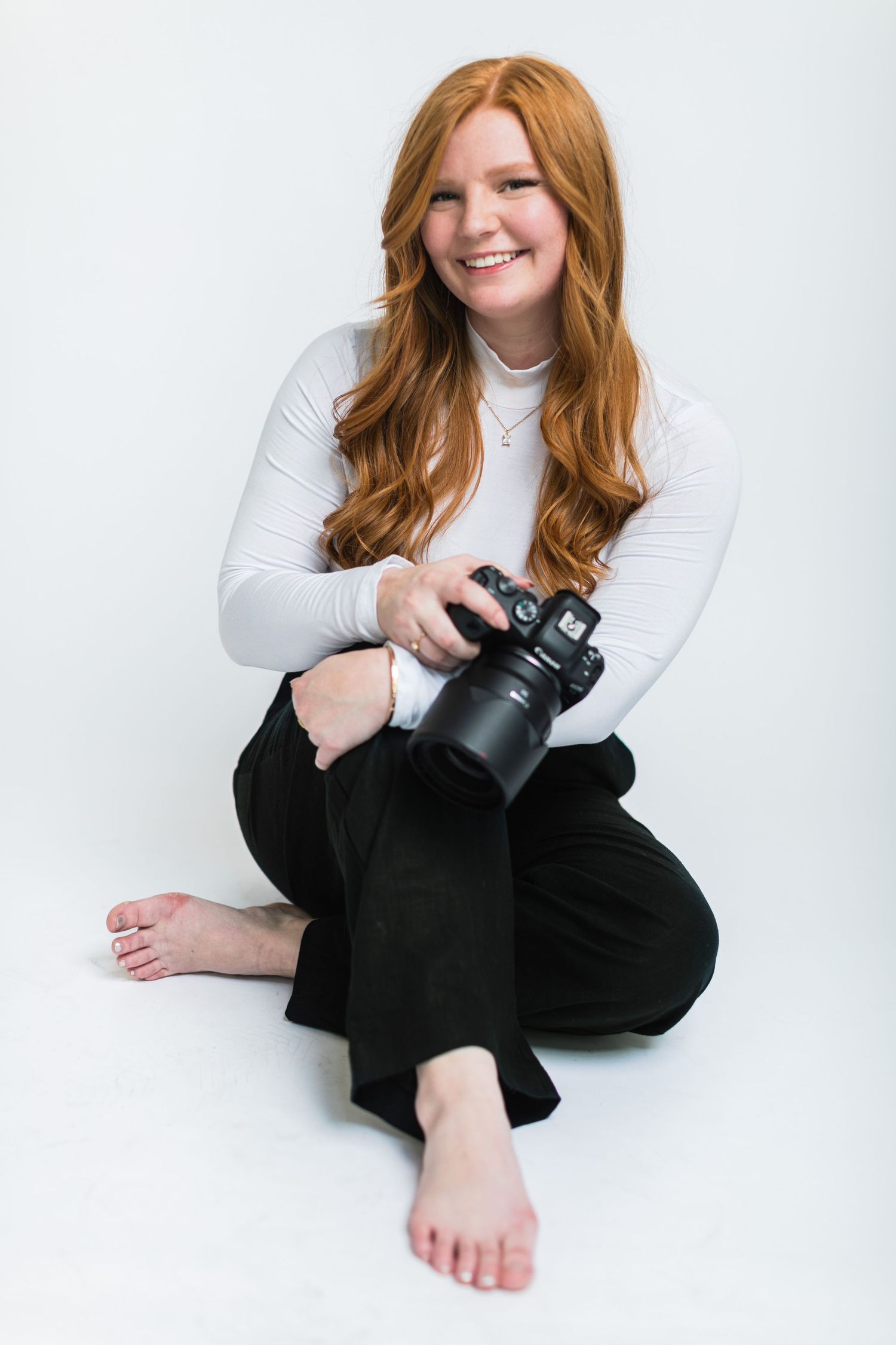 Smiling redhead female photographer sits cross-legged holding a professional camera on white background.