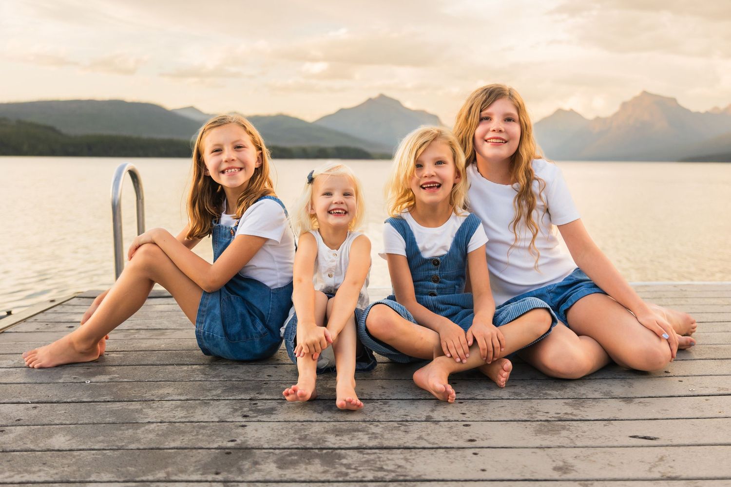 Four children sitting on a dock by a mountain lake, smiling and laughing together.