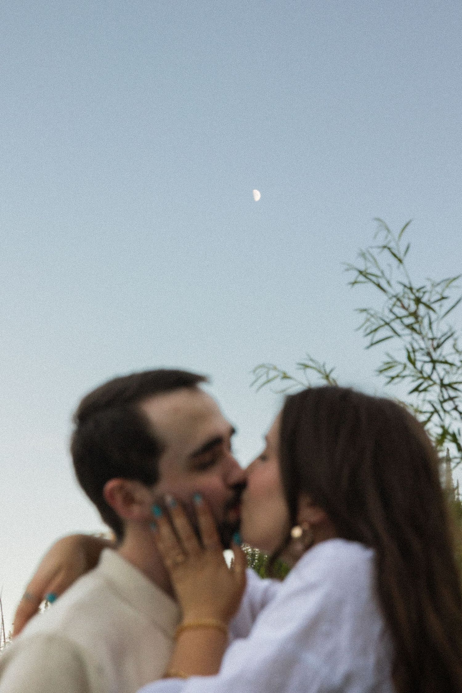 Couple sharing a romantic kiss outdoors under a crescent moon in soft evening light.