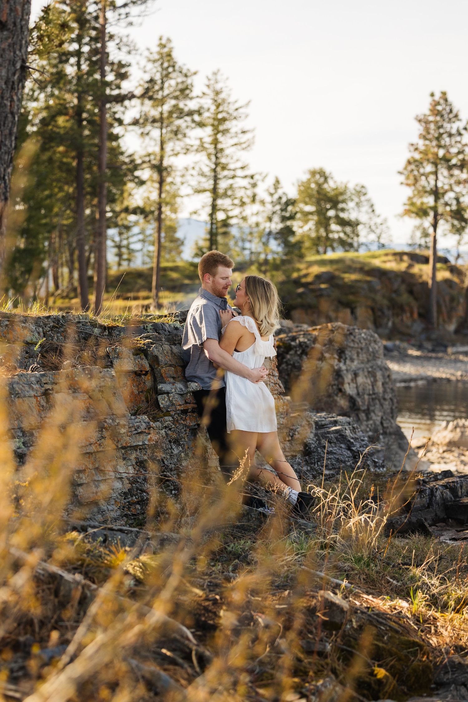 Romantic couple embracing against stone wall in golden sunset light among pine trees outdoors.