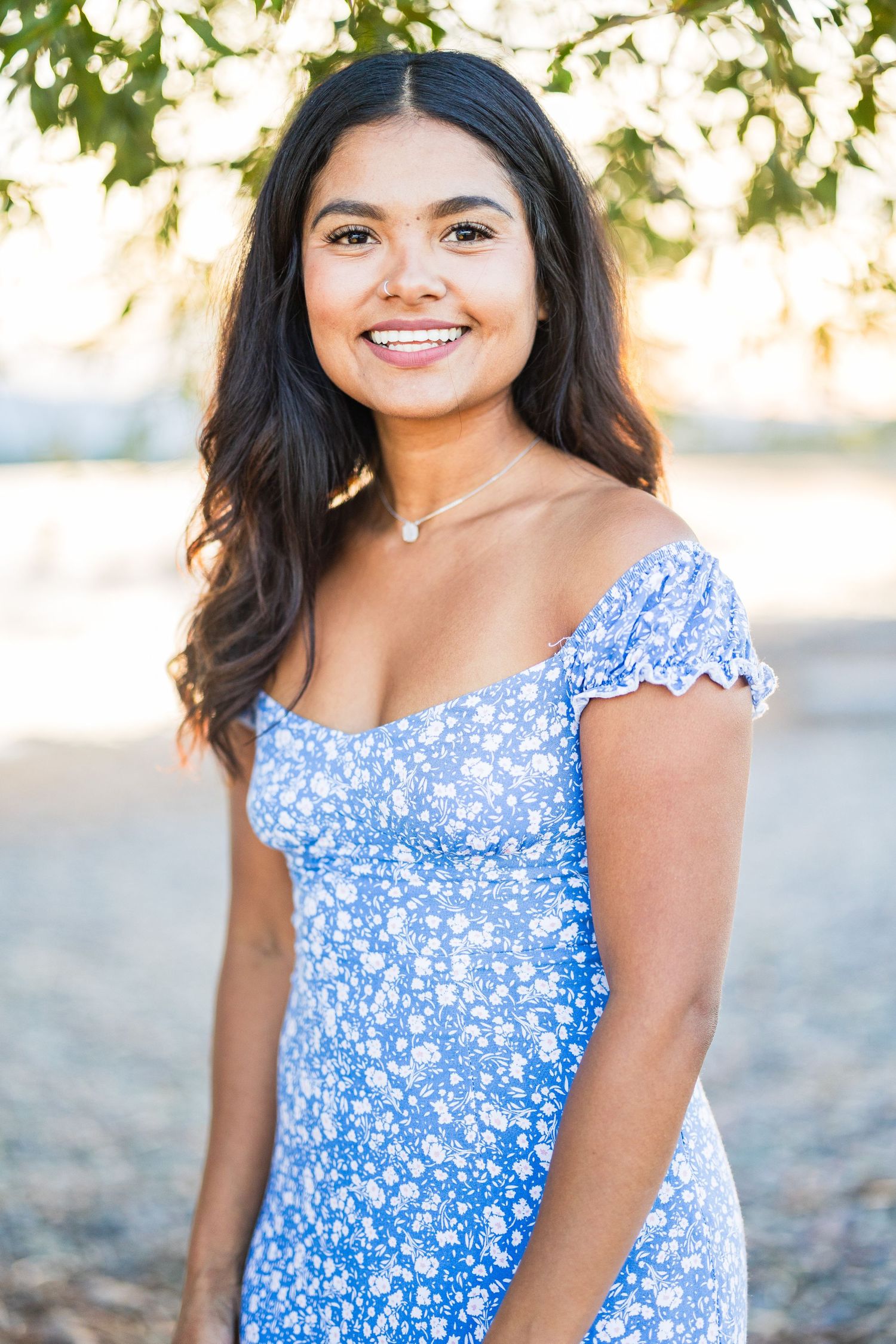Young woman in a blue floral off-shoulder dress smiling outdoors under leafy trees.