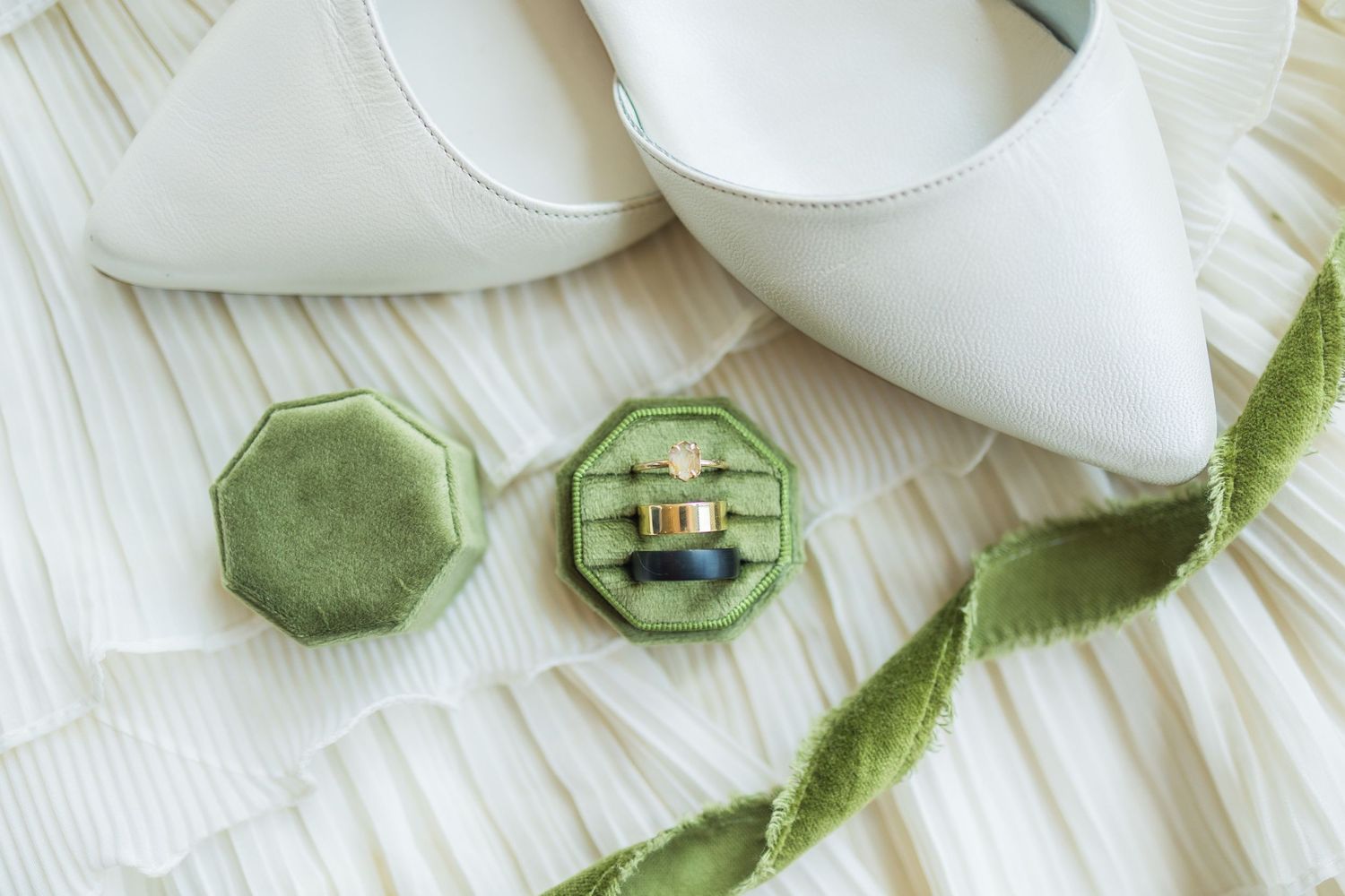 Green velvet octagonal ring box with wedding bands beside white bridal heels on pleated fabric.