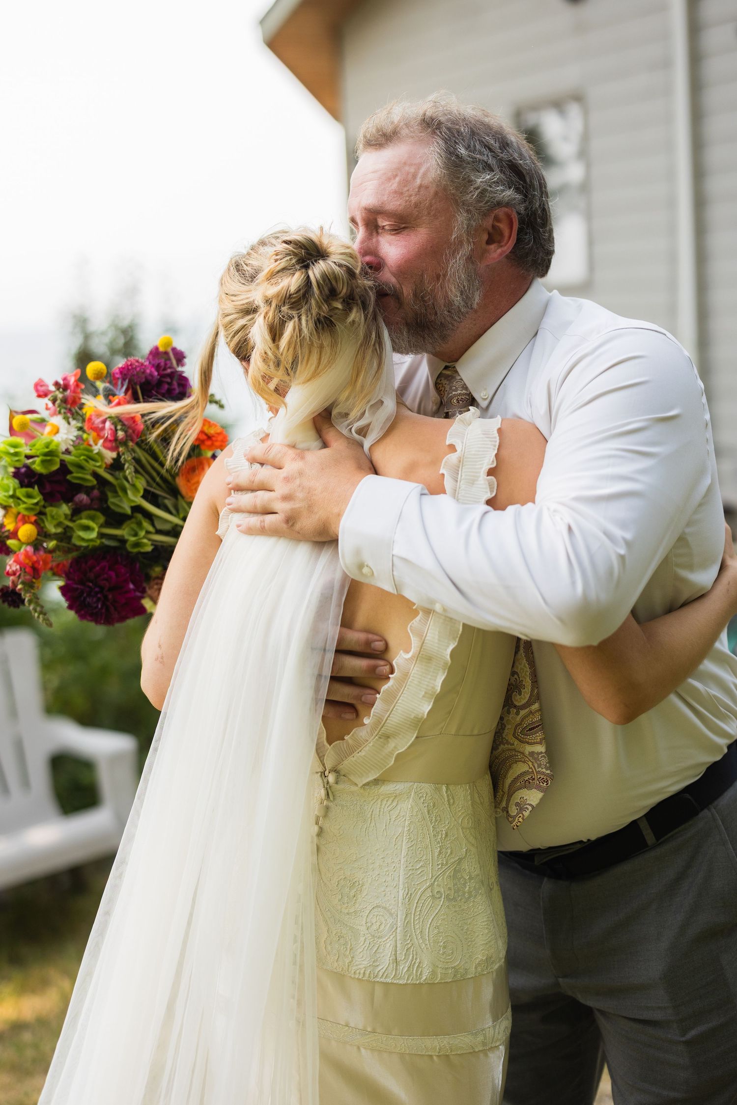 Father kissing bride on the head in an emotional wedding day hug, bride holding colorful bouquet outdoors.