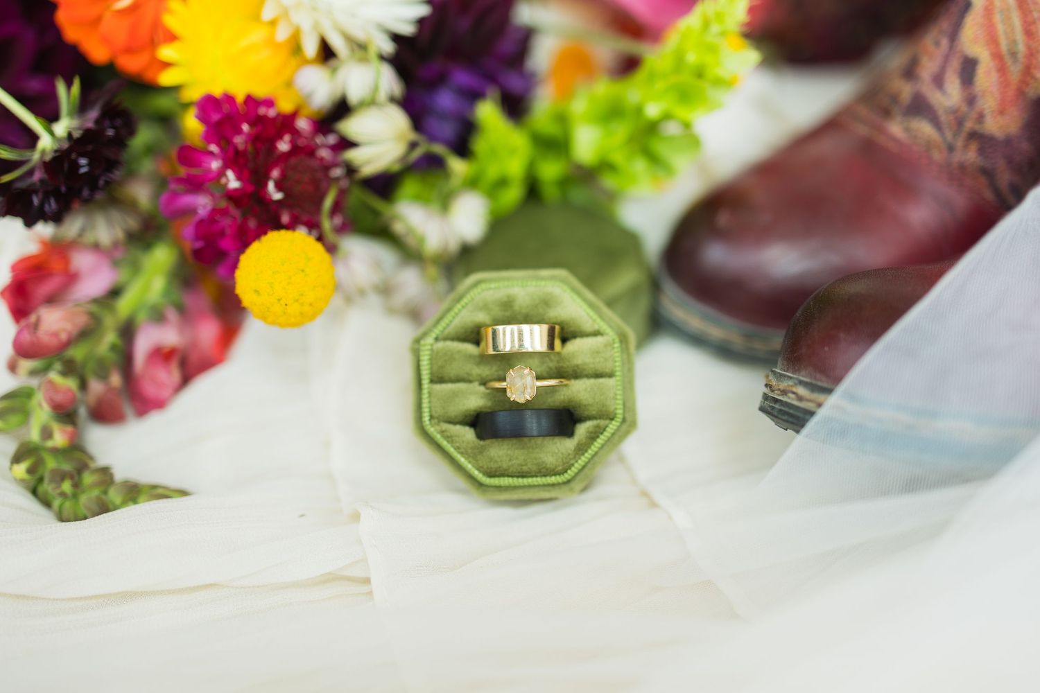Gold wedding ring displayed in a green velvet octagonal ring box, surrounded by colorful wildflowers and a bridal veil.