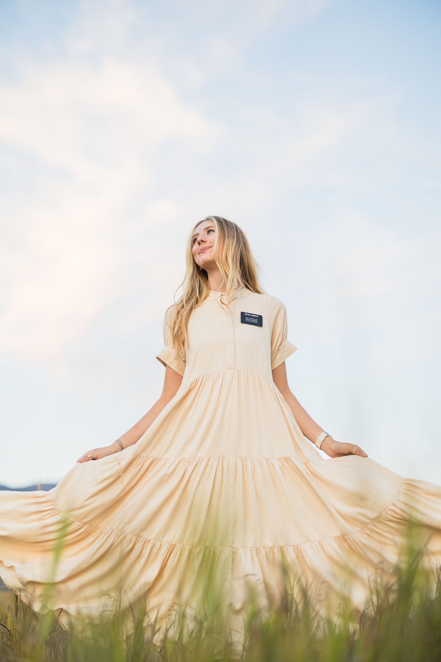 Blonde woman in cream missionary dress twirling in a field under a bright sky.