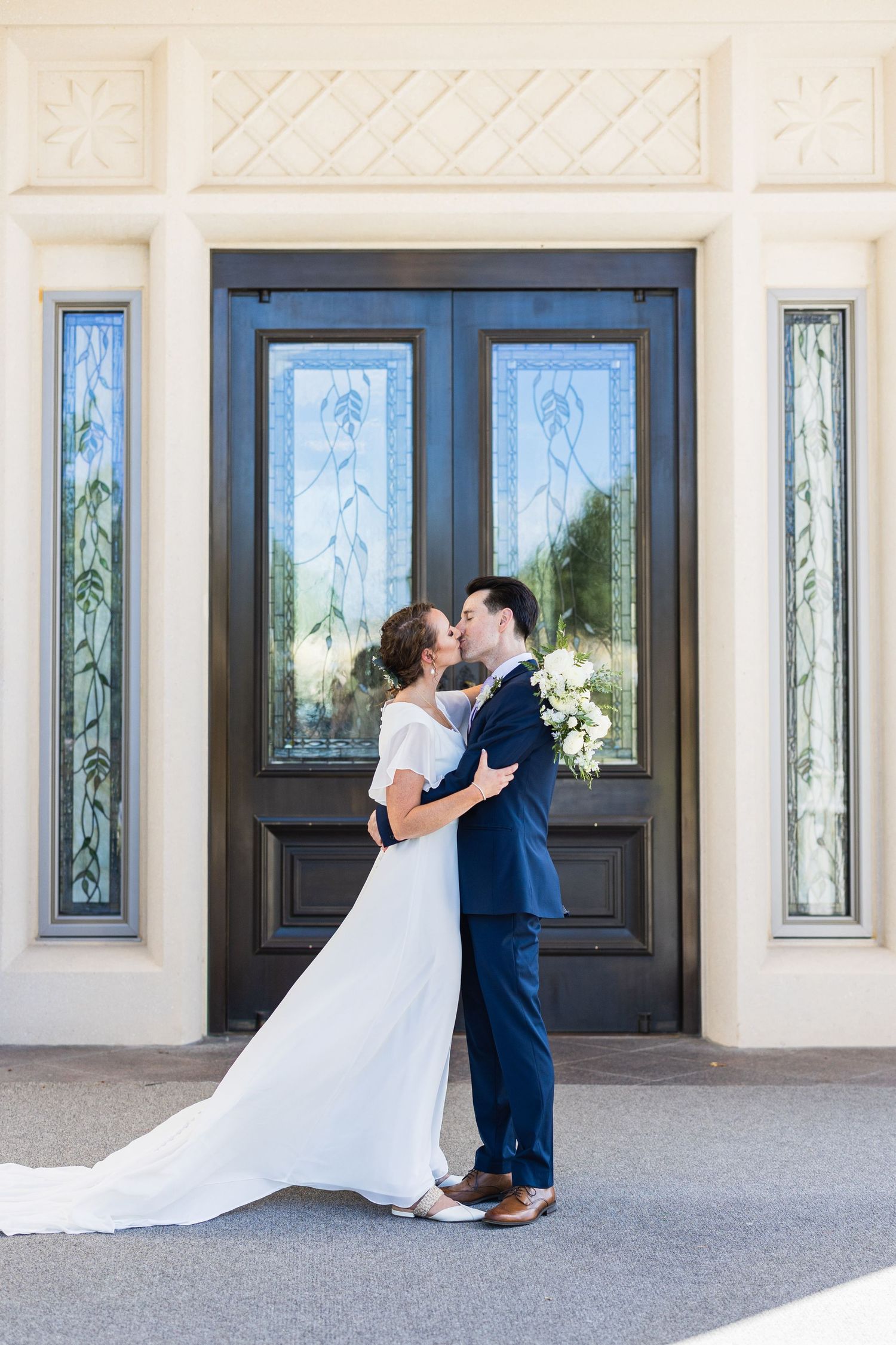 Bride and groom share a kiss in front of elegant double doors with stained glass windows.