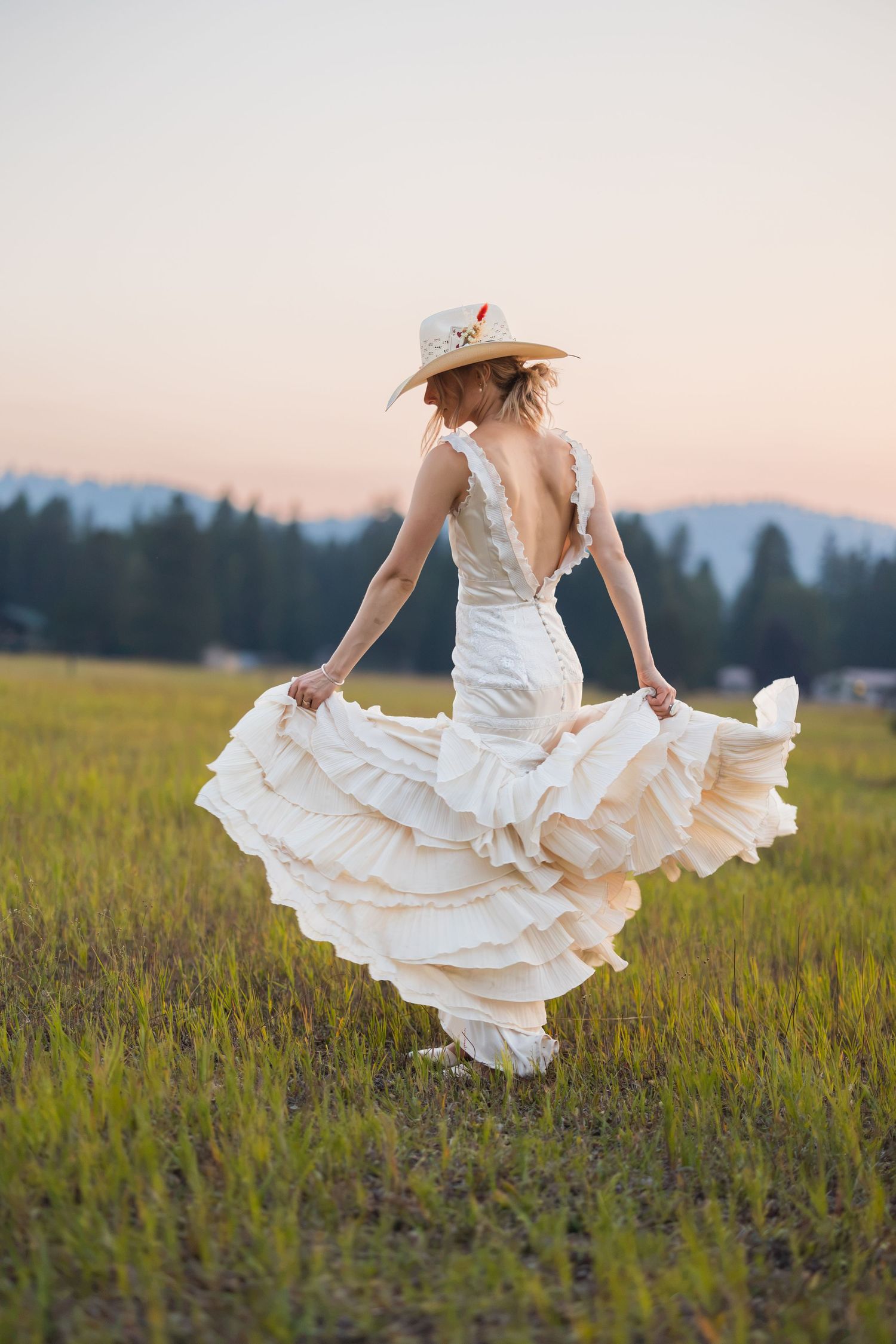 Woman in white ruffled backless dress and straw hat twirling in green meadow at sunset with mountains behind.