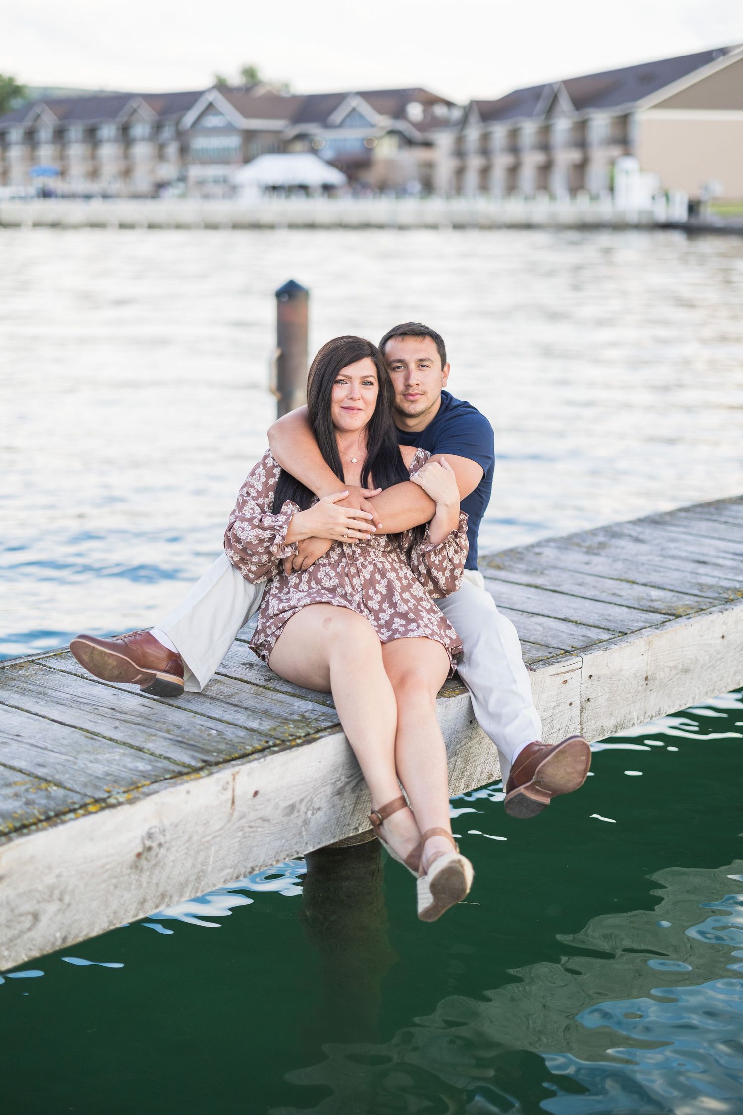 Couple sits closely together on waterfront dock embracing at sunset with marina in background.
