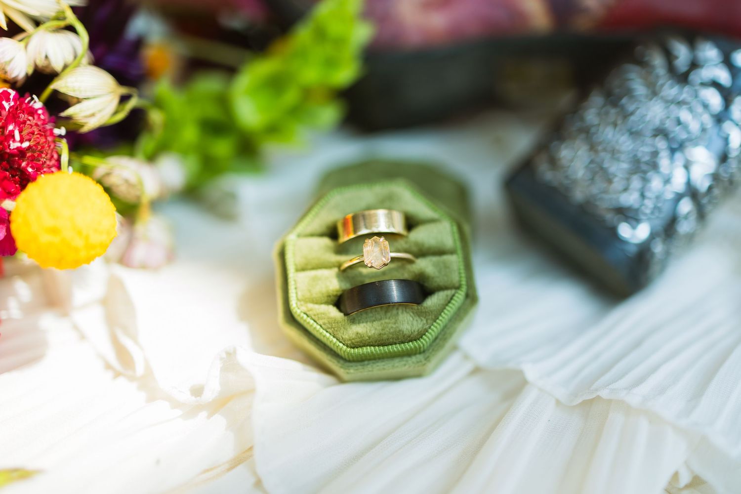 Green velvet ring box holding two wedding bands and a diamond engagement ring, surrounded by colorful flowers and accessories.