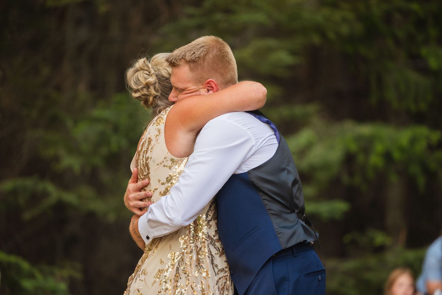 Bride in gold embroidered gown and groom in blue vest share an emotional embrace during outdoor wedding reception.