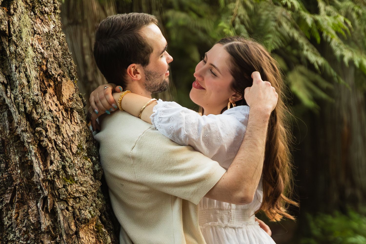 Couple embracing romantically by a tree in a lush green forest, woman in white dress smiling up at man.