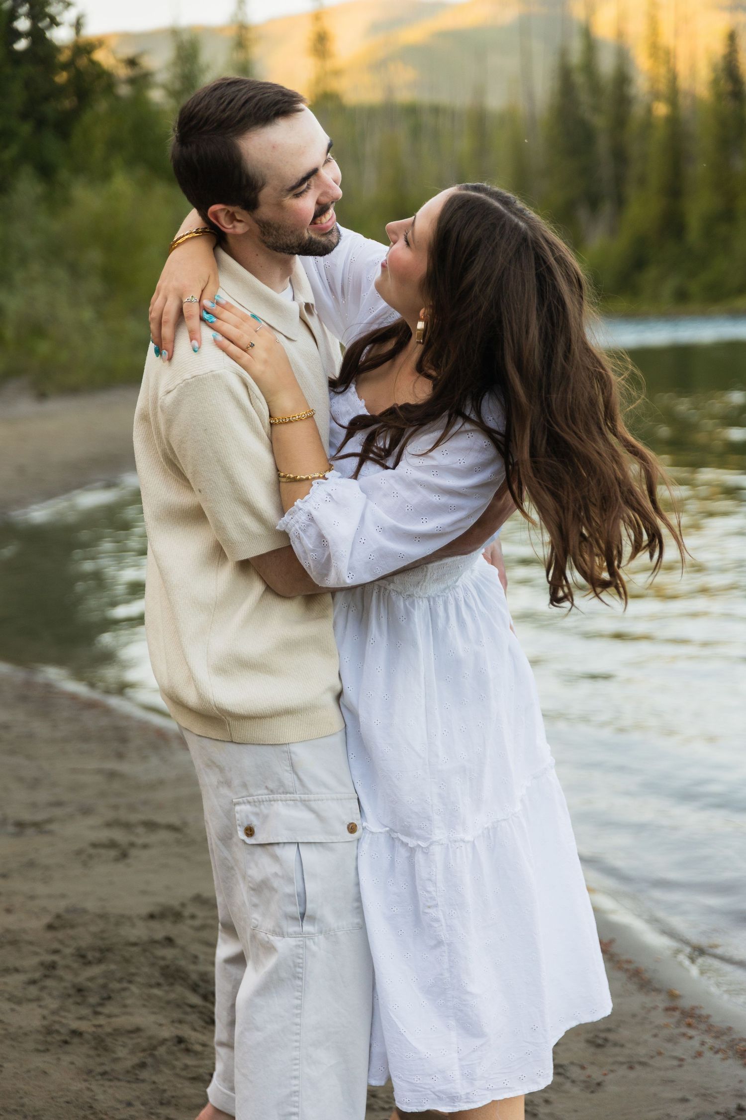 A couple embraces and laughs together on a lakeside beach surrounded by mountains at golden hour.
