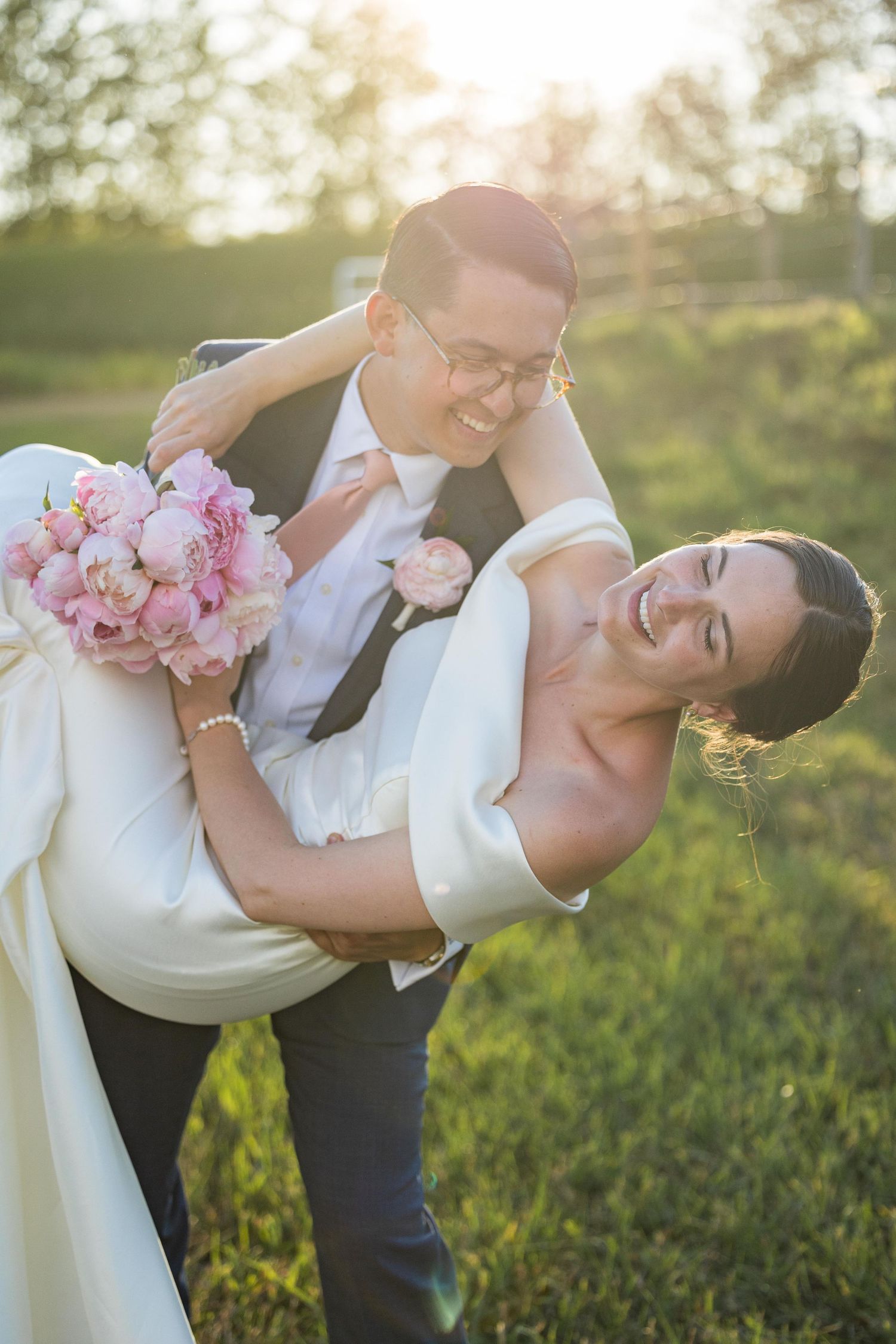Joyful groom lifting laughing bride holding pink peony bouquet in sunlit green field.