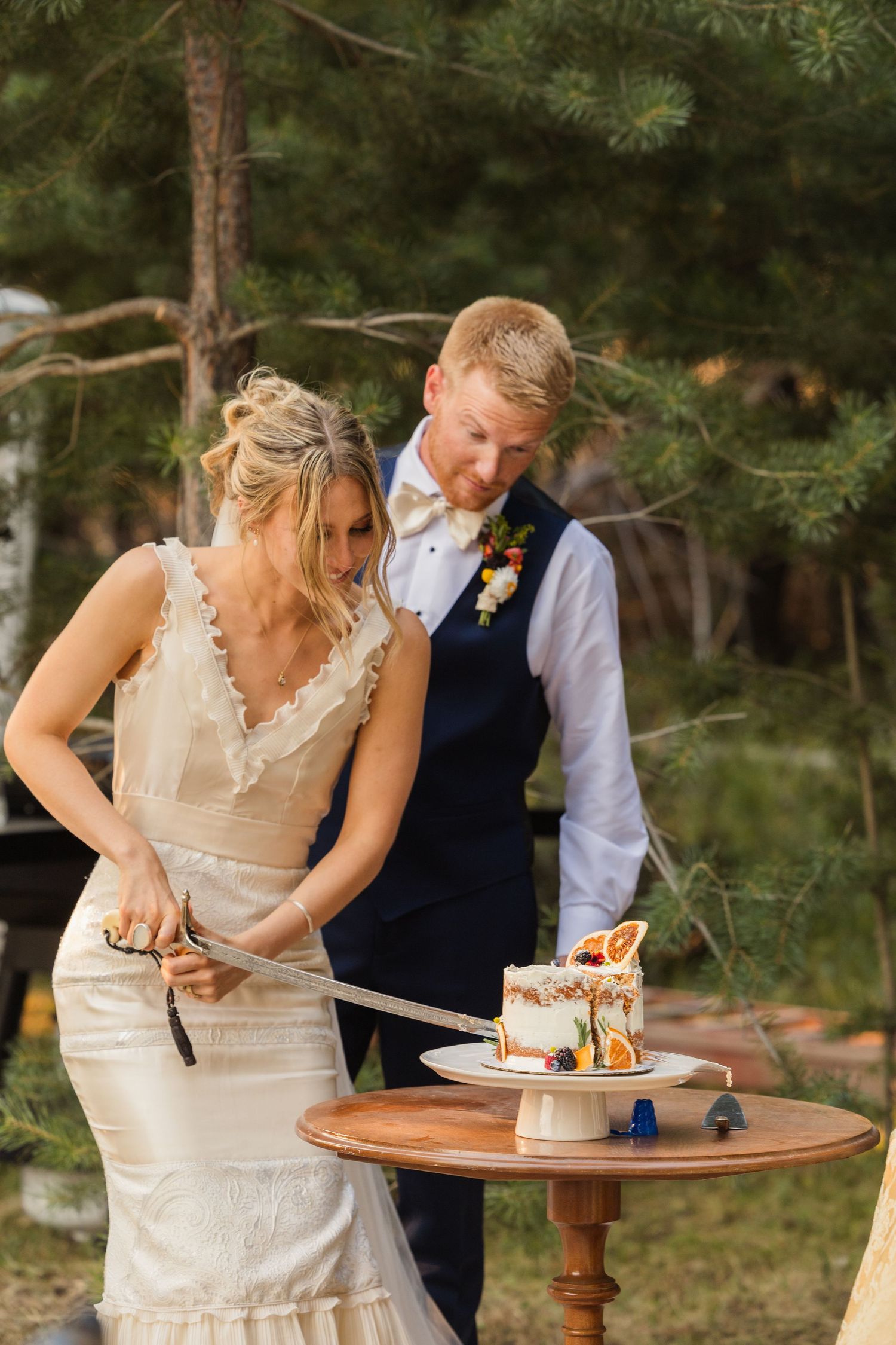 Bride and groom cutting wedding cake with a sword at an outdoor forest wedding ceremony.