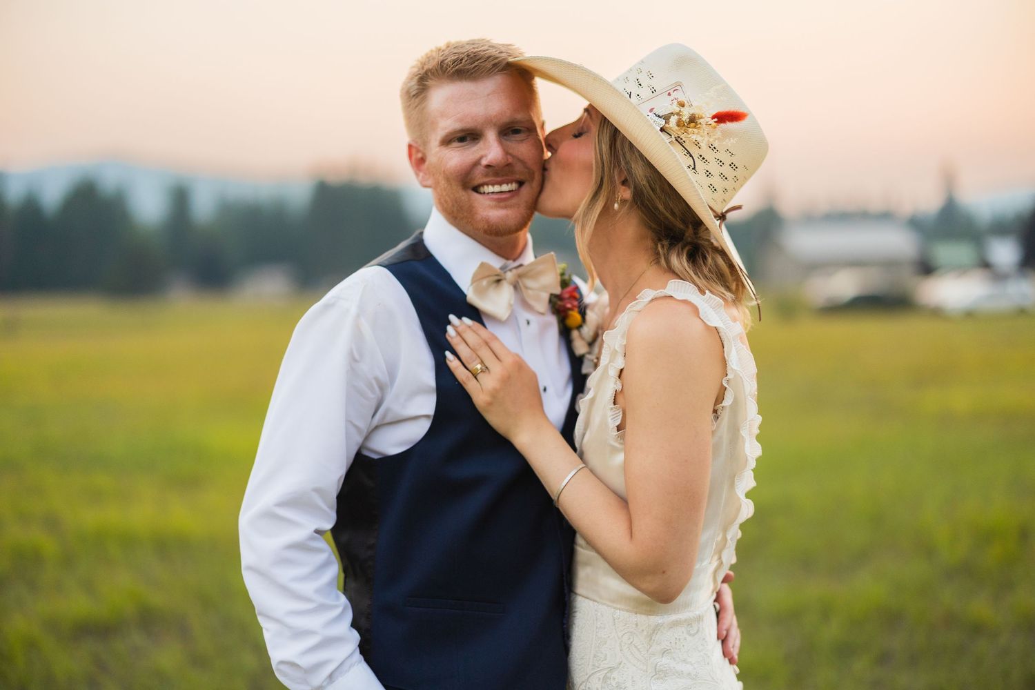 Bride in lace dress and cowboy hat kisses groom in navy vest during golden hour outdoor wedding portrait.