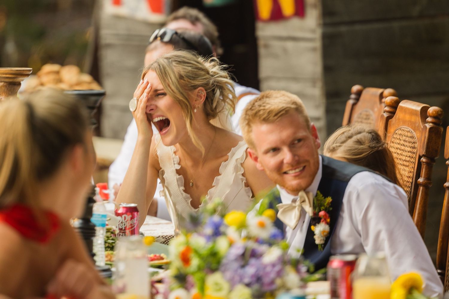 Bride laughing joyfully with groom smiling beside her at a colorful outdoor wedding reception dinner table.