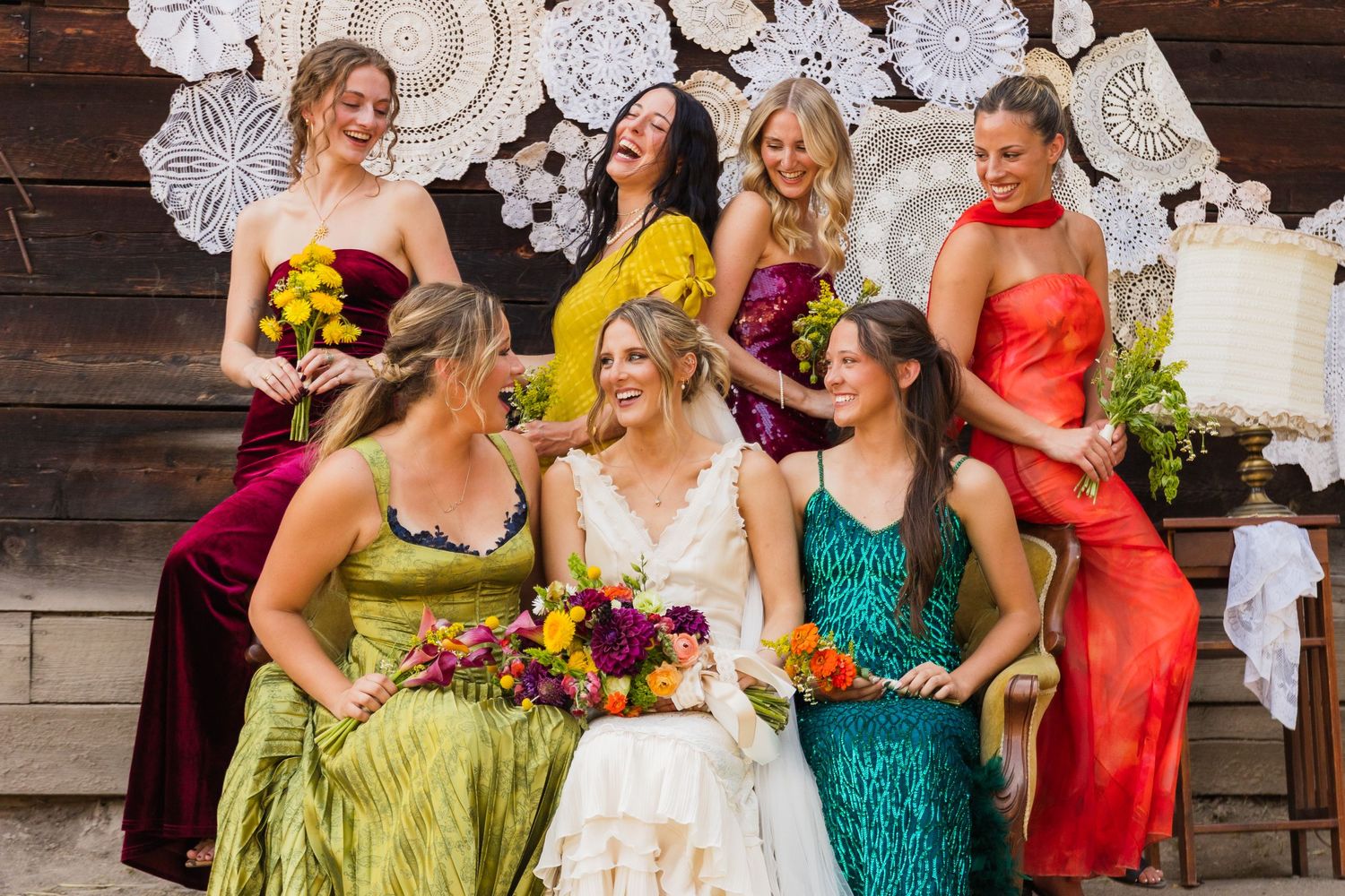 Bride in white gown surrounded by bridesmaids in colorful dresses in front of a rustic barn wall.
