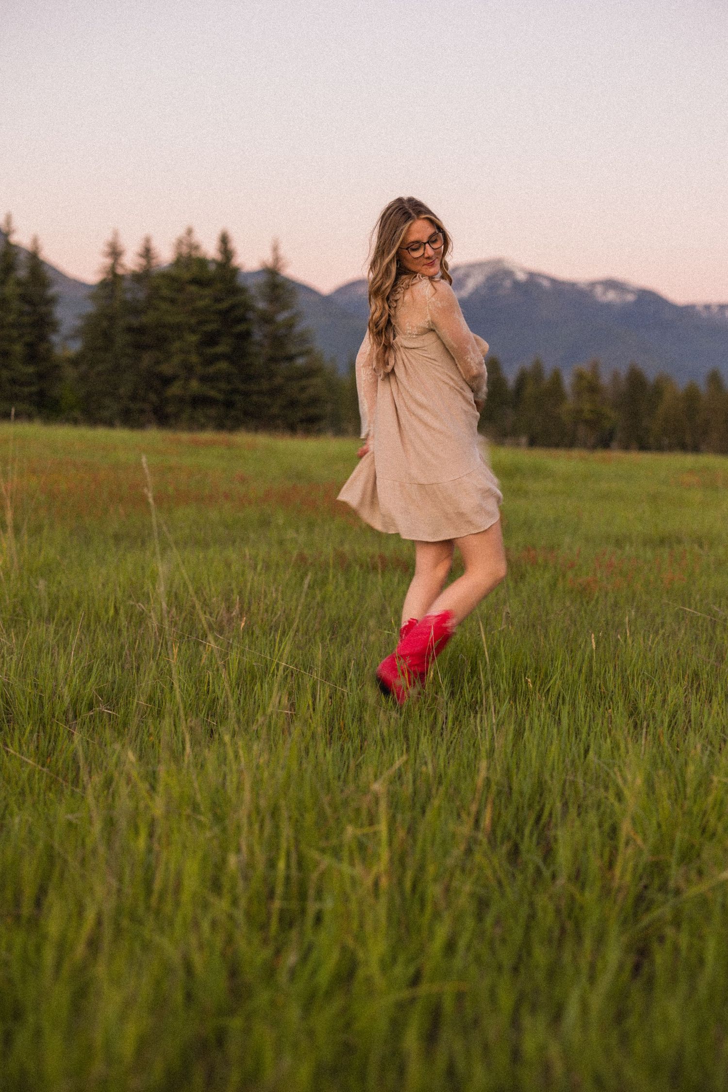 Young woman in beige dress and red cowboy boots walking in a mountain meadow at dusk.