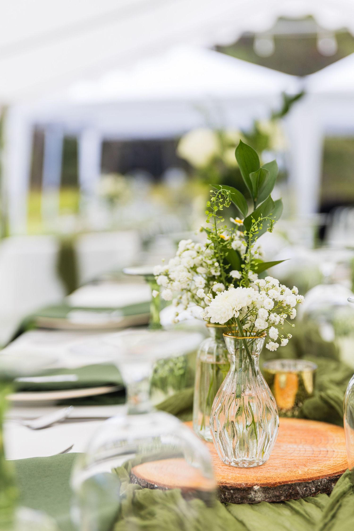 Elegant wedding table centerpiece with white flowers in glass vase on wood slice with green runner.