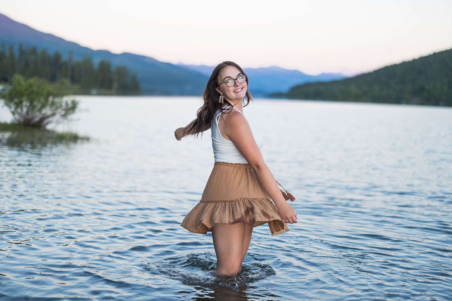 Young woman in white top and tan skirt twirling in a mountain lake at sunset.