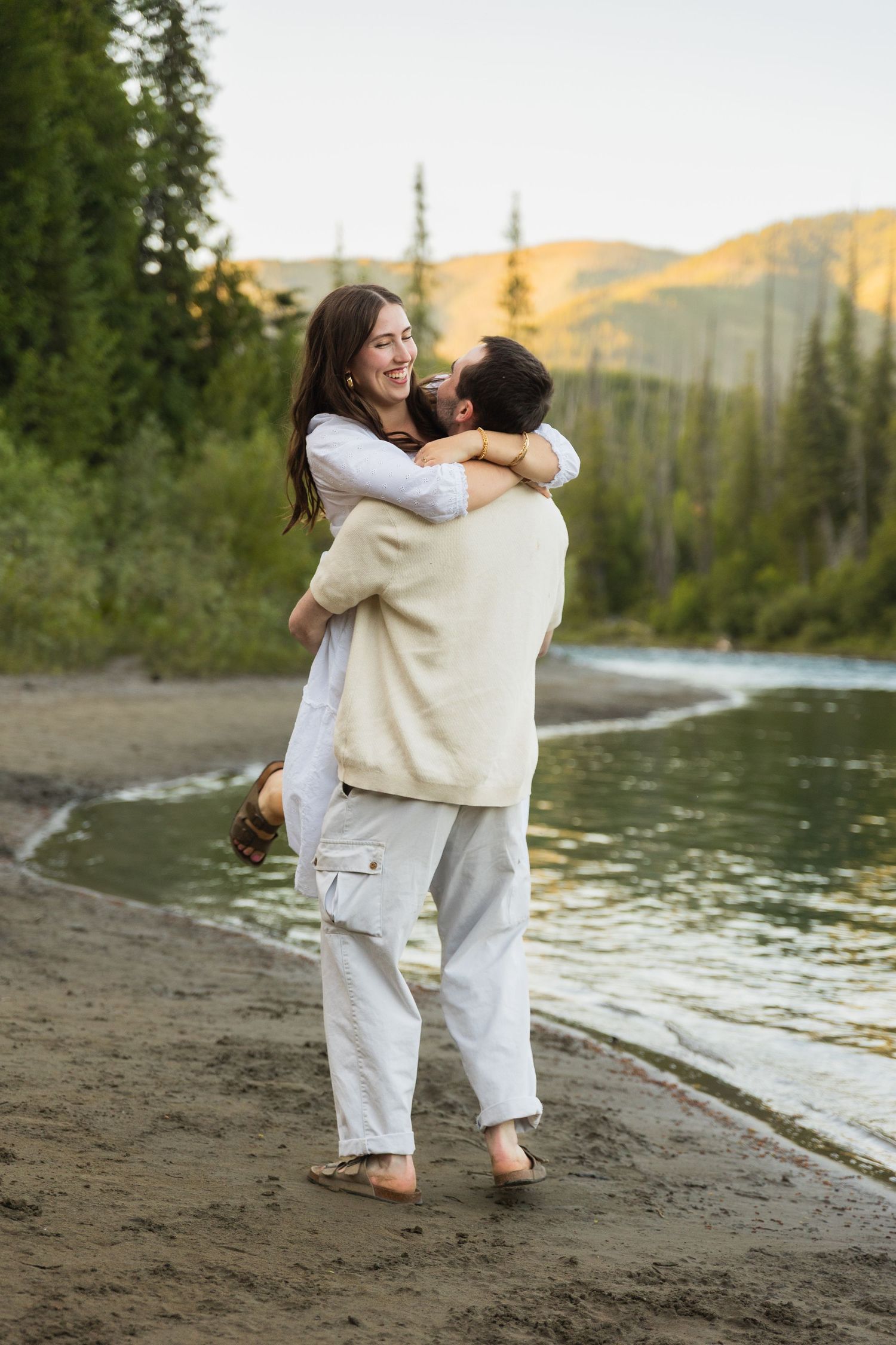 Man joyfully lifting woman on sandy riverbank surrounded by pine trees at sunset.
