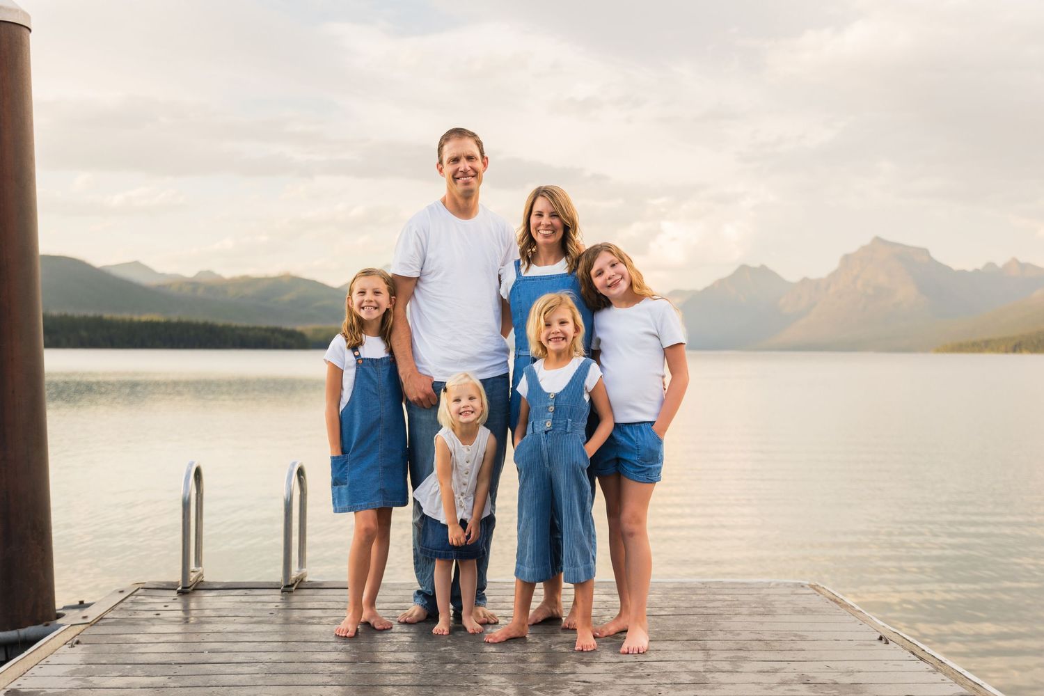Family of six smiling on a dock with a mountain lake and cloudy sky behind them.