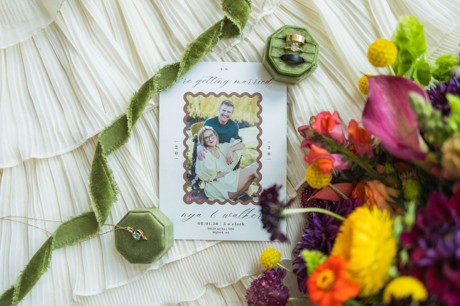 Wedding flat lay with save the date card, velvet ring box, colorful floral bouquet, and ribbon on white fabric.