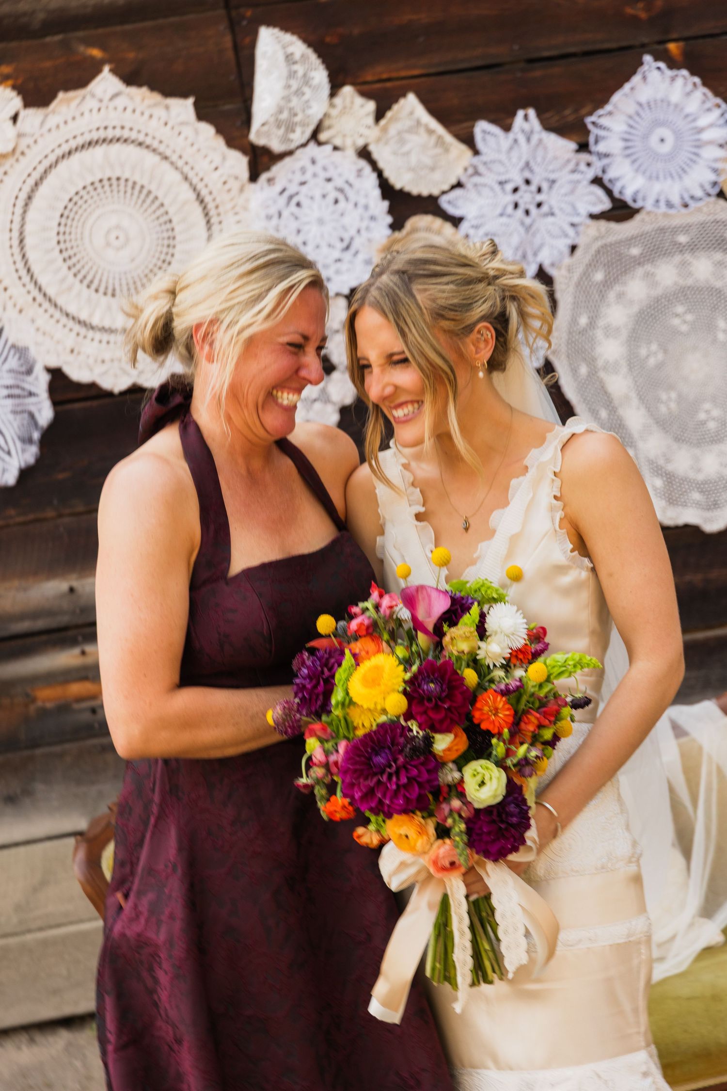Two women laughing together at a wedding, one in a white bridal gown holding a vibrant colorful bouquet, with lace doilies backdrop.