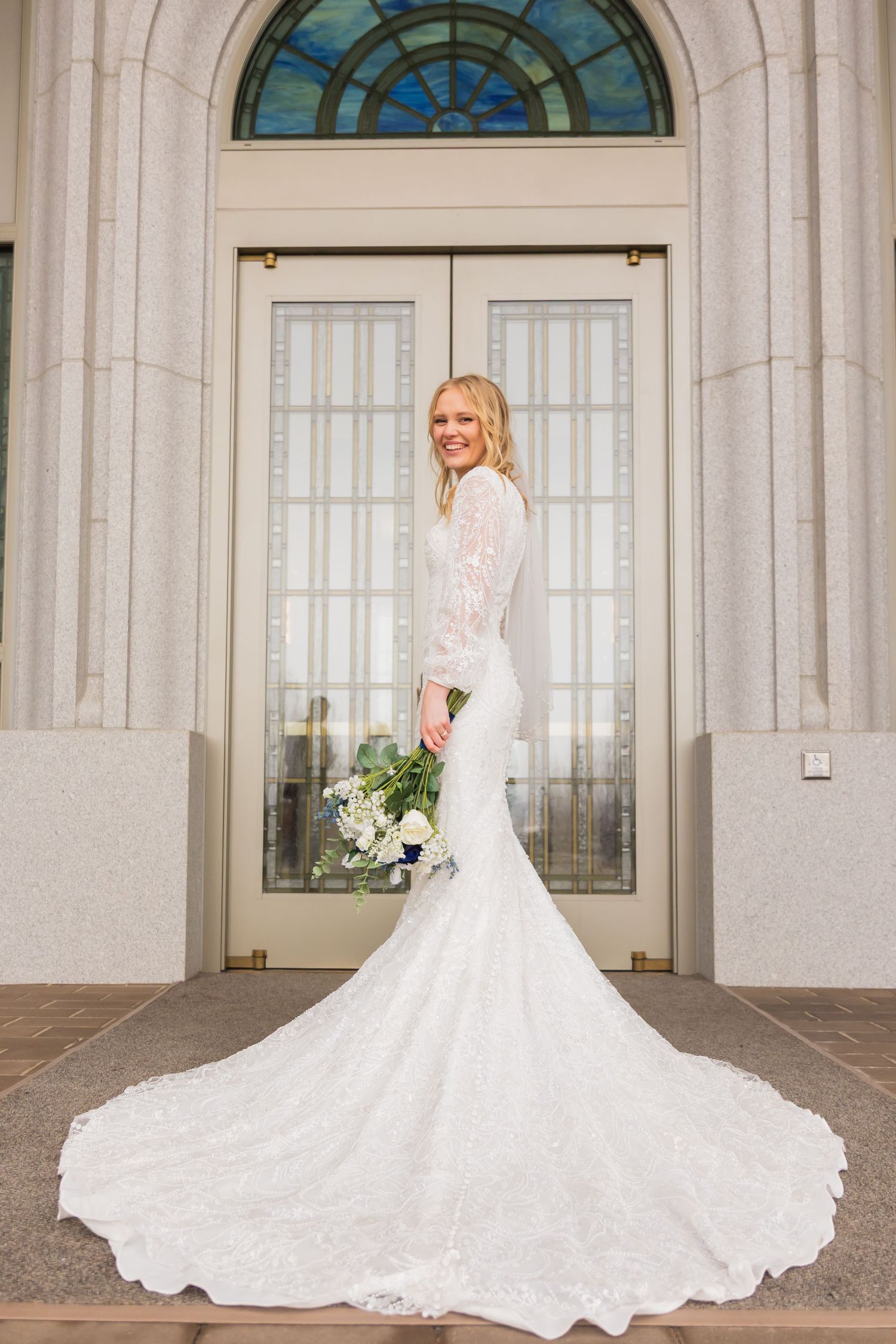 Elegant bride in long-sleeve lace mermaid gown with cathedral train poses outside ornate temple doors.