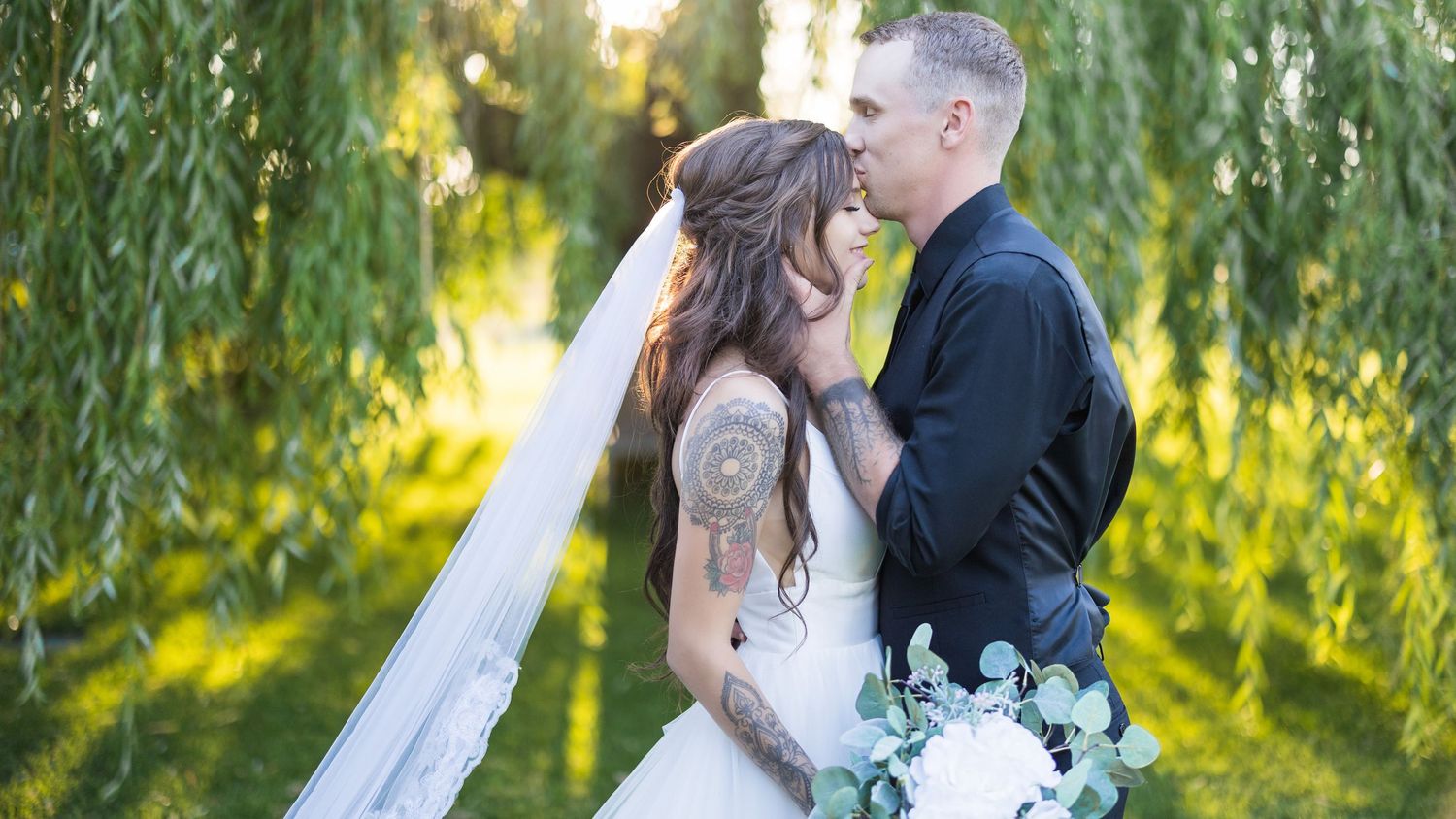 Bride with tattoos and groom share an intimate moment under lush green weeping willow trees.